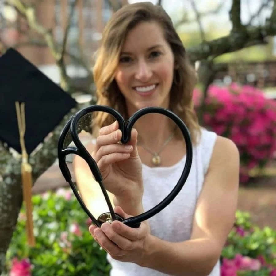 Nurse Graduation 桌布 壁紙 — 一位微笑的畢業生在花園中手持心形聽診器，旁邊放著畢業帽。