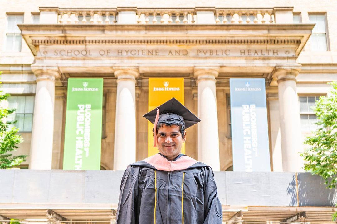 college 桌布 壁紙 — 一位微笑的畢業生身穿畢業袍和學士帽，在晴朗的日子裡自豪地站在School of Hygiene and Public Health大樓前。