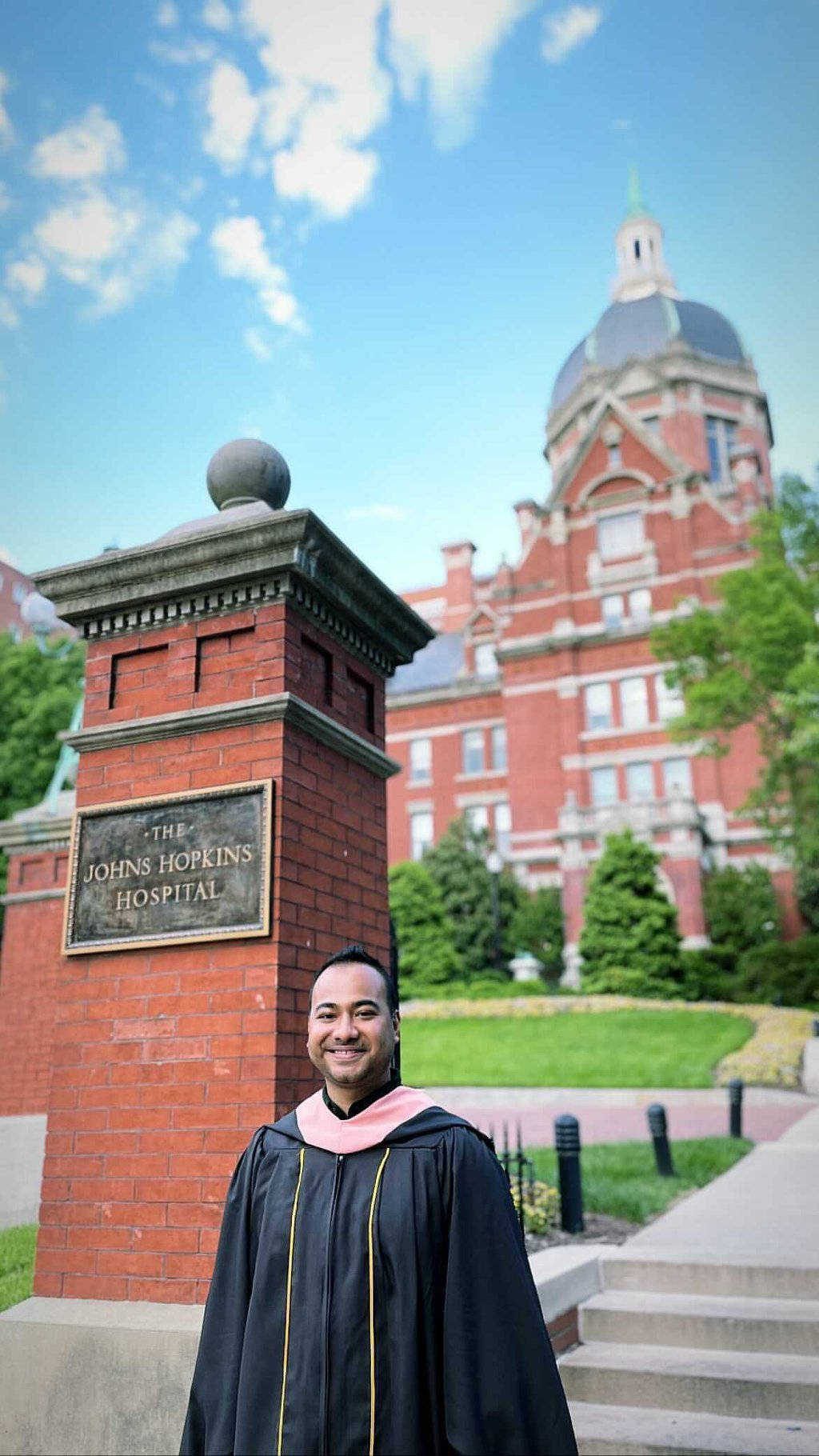 college 桌布 壁紙 — 一位微笑的畢業生身穿黑色畢業袍和粉紅色披肩，自豪地站在寫有約翰霍普金斯醫院的磚柱前，背景是明亮的藍天。