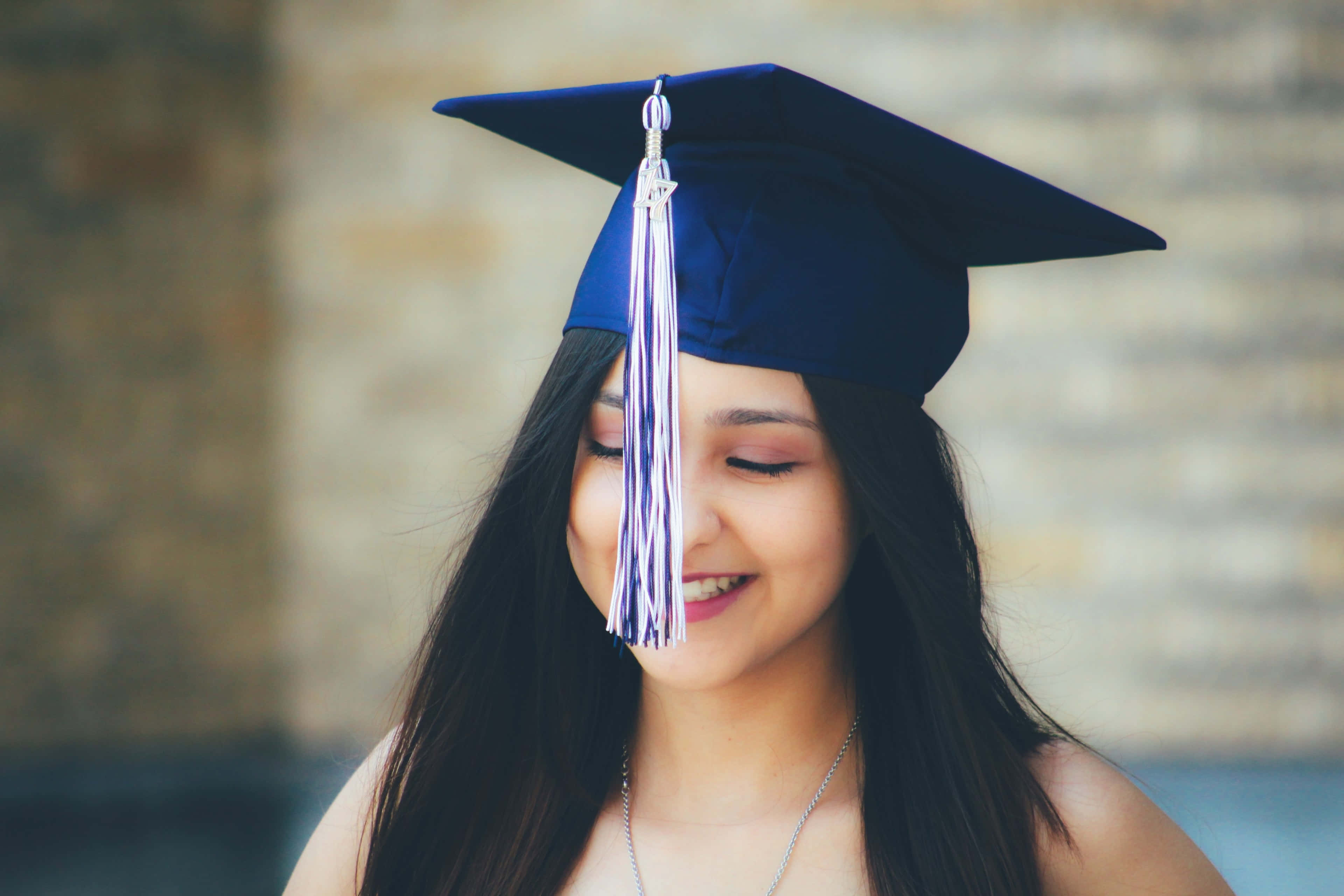 Graduation Ceremony 桌布 壁紙 — 一位微笑的畢業生閉著眼睛，戴著藍色學士帽和流蘇，背景是模糊的磚牆。