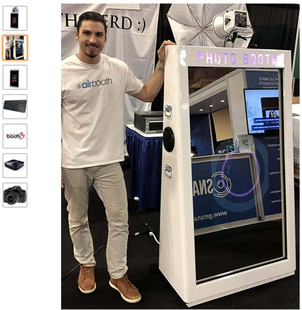 Photo Booth 桌布 壁紙 — 一位微笑的白T恤男子站在現代白色照相亭旁，活動現場有工作室燈光和品牌背景。