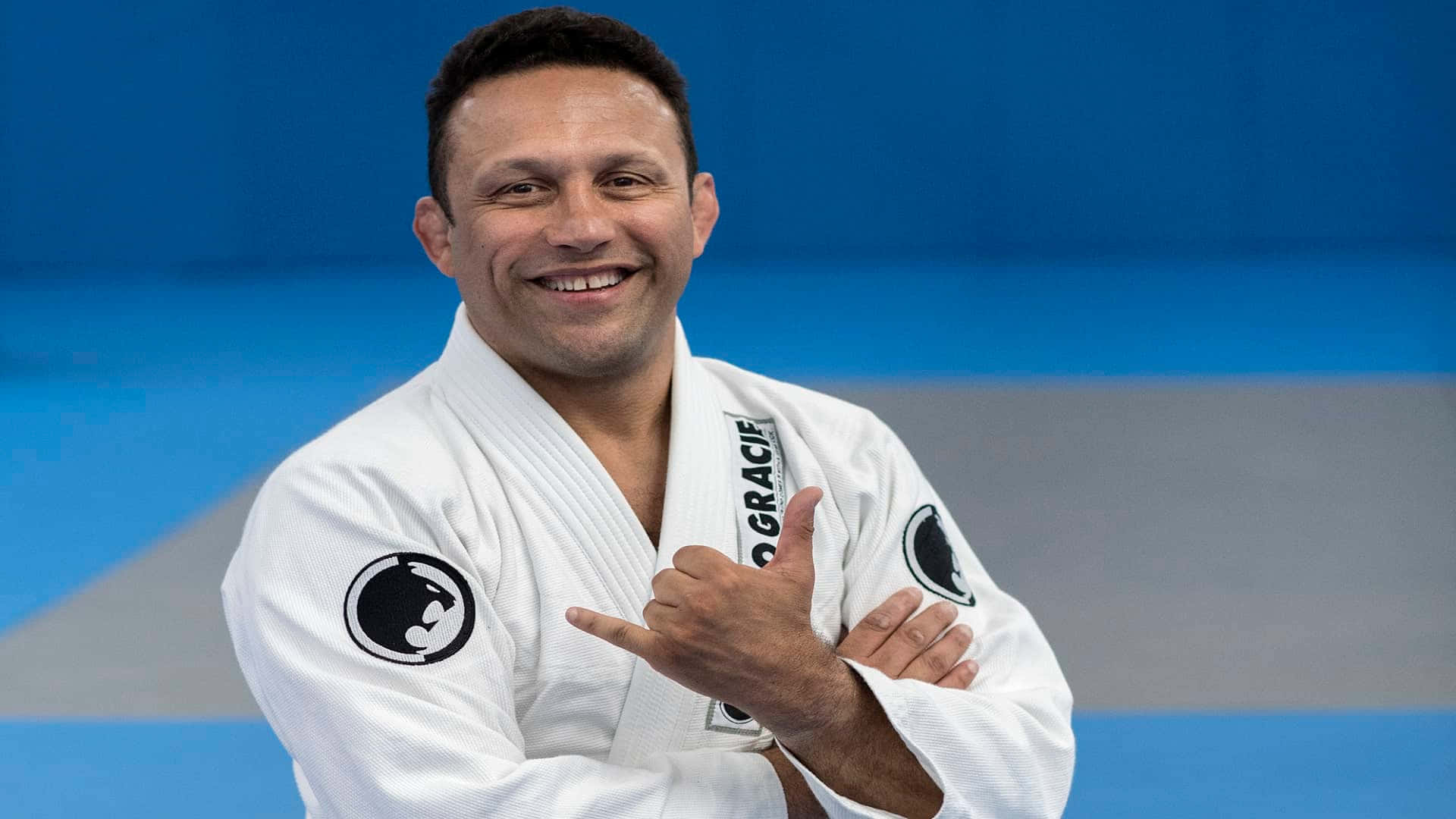martial arts 桌布 壁紙 — 一位微笑的白衣柔道男子在藍灰色墊子背景下豎起大拇指。