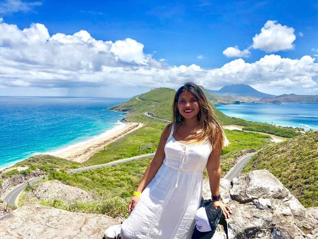 Vacation 桌布 壁紙 — 一位微笑的白裙女士在岩石懸崖上擺姿勢，俯瞰著充滿活力的碧綠海洋和蜿蜒的海岸公路，天空湛藍，白雲朵朵。