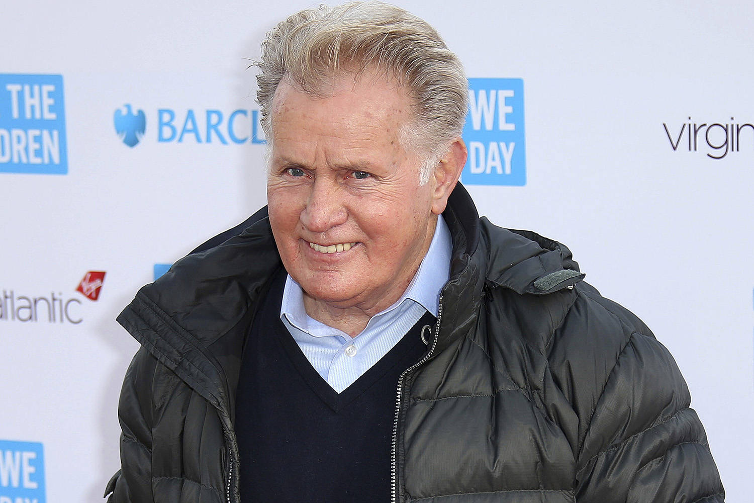 Celebrity 桌布 壁紙 — 一位微笑的白髮年長男士在品牌活動背景前，穿著黑色羽絨外套，內搭藍色襯衫和深色毛衣。