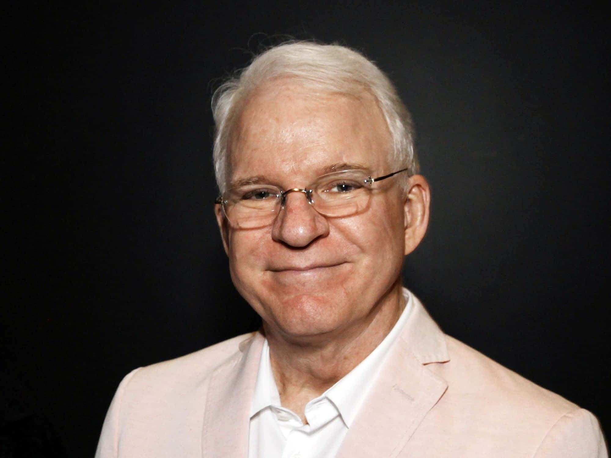 steve martin 桌布 壁紙 — 一位微笑的白髮老人戴著眼鏡，身穿淺粉色西裝外套，背景為深色。
