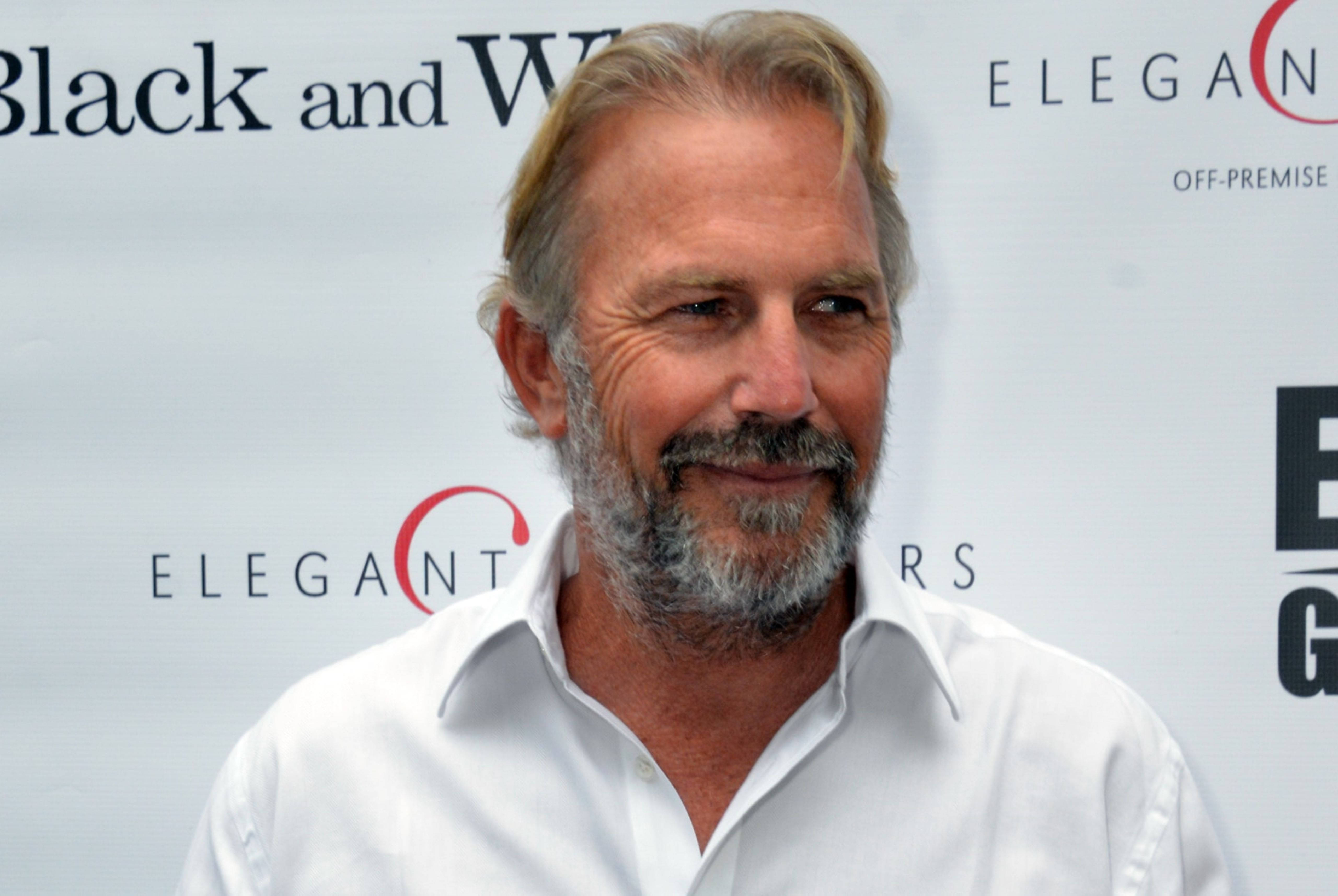 Kevin Costner 桌布 壁紙 — 一位微笑的白髮鬍鬚男士穿著白襯衫，站在品牌活動背景前。