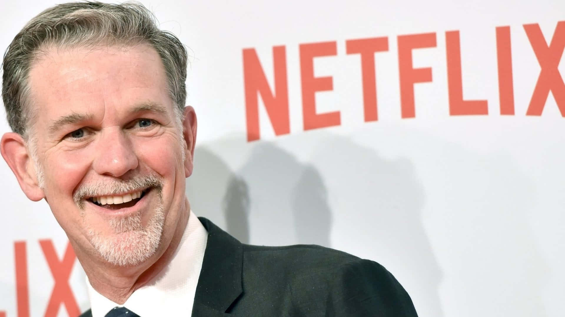 Netflix 桌布 壁紙 — 一位微笑的白髮鬍鬚男士身穿黑色西裝，站在白色背景前，背景有紅色Netflix標誌。