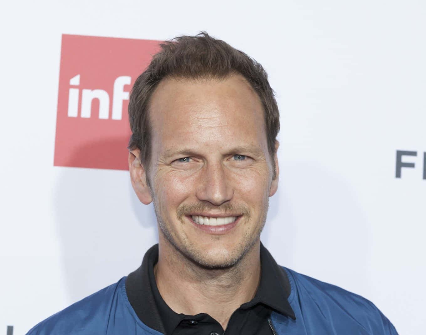 Patrick Wilson 桌布 壁紙 — 一位微笑的短棕髮藍眼男士，身穿藍色外套內搭黑色襯衫，背景為白色並帶有紅色和黑色標誌。