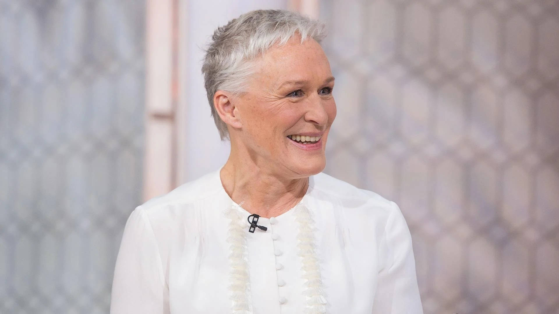 Actress HD 桌布 壁紙 — 一位微笑的短灰髮女性穿著白色襯衫，前面有裝飾性設計，背景為圖案背景。