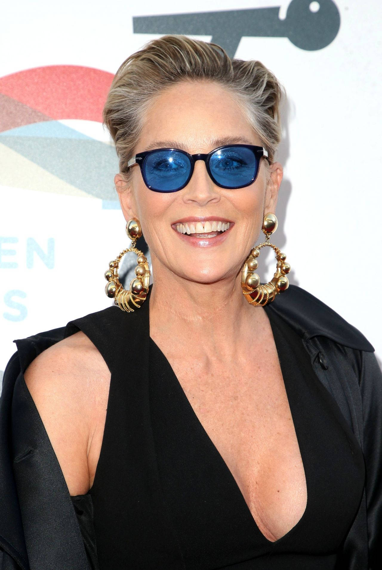 Celebrity 桌布 壁紙 — 一位微笑的短金髮女性，戴著大膽的藍色太陽眼鏡和大金環耳環，出席紅地毯活動。