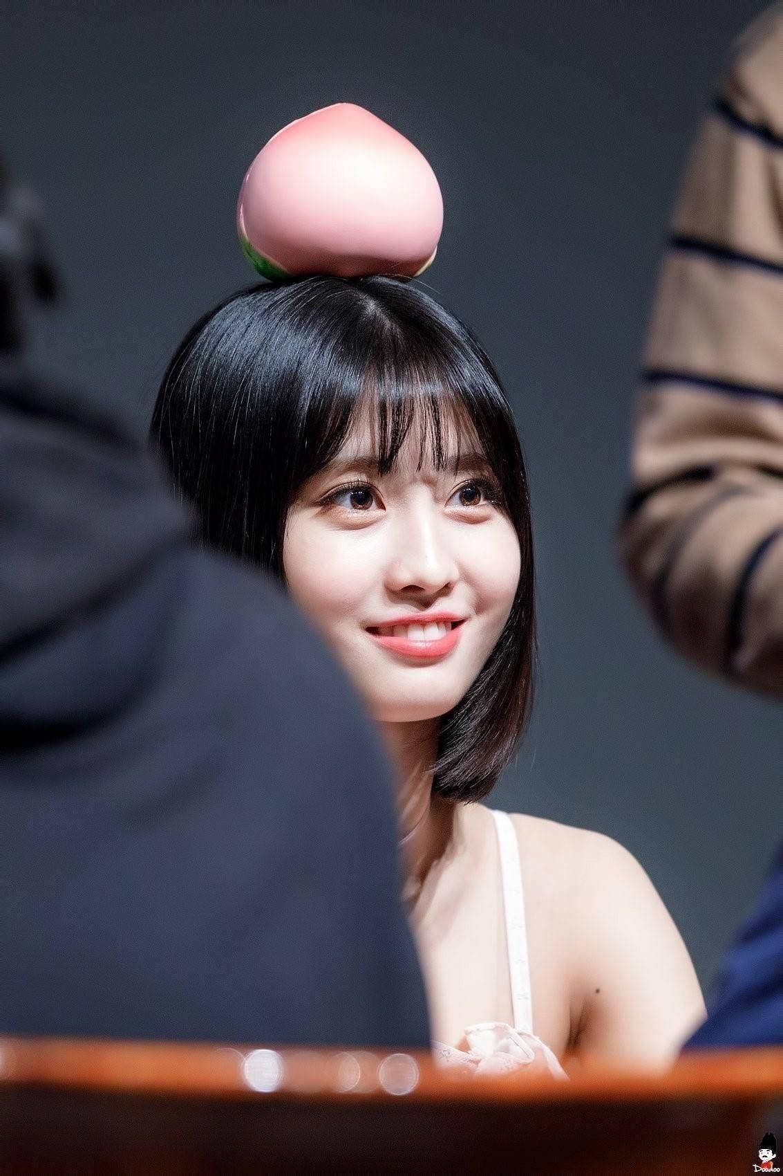 Momo 桌布 壁紙 — 一位微笑的短黑髮年輕女性在室內坐著時，將一個粉紅色的桃子平衡在頭上。