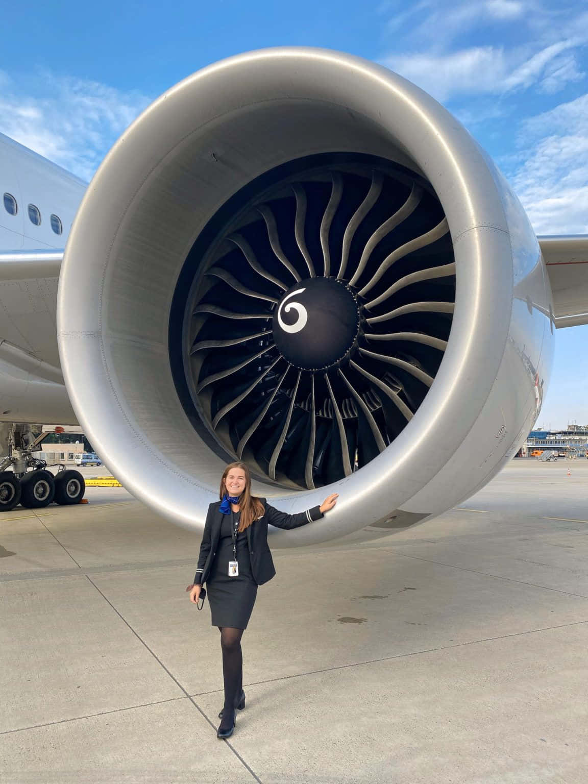 Aircraft 桌布 壁紙 — 一位微笑的空服員在陽光明媚的機場停機坪上，自信地站在巨大的飛機引擎旁邊，背景是藍天。