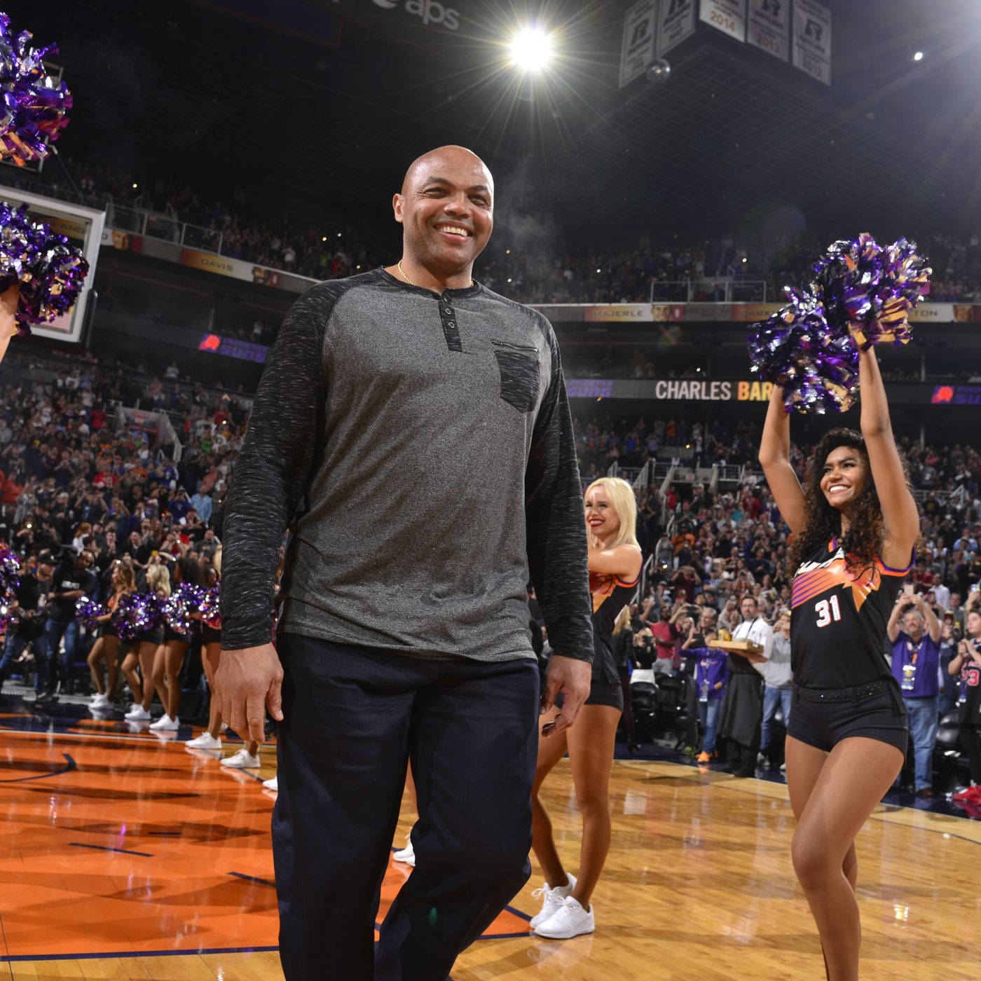 NBA 桌布 壁紙 — 一位微笑的籃球教練自信地走過燈光明亮的球場，兩旁是歡呼的啦啦隊和擠滿觀眾的體育場。