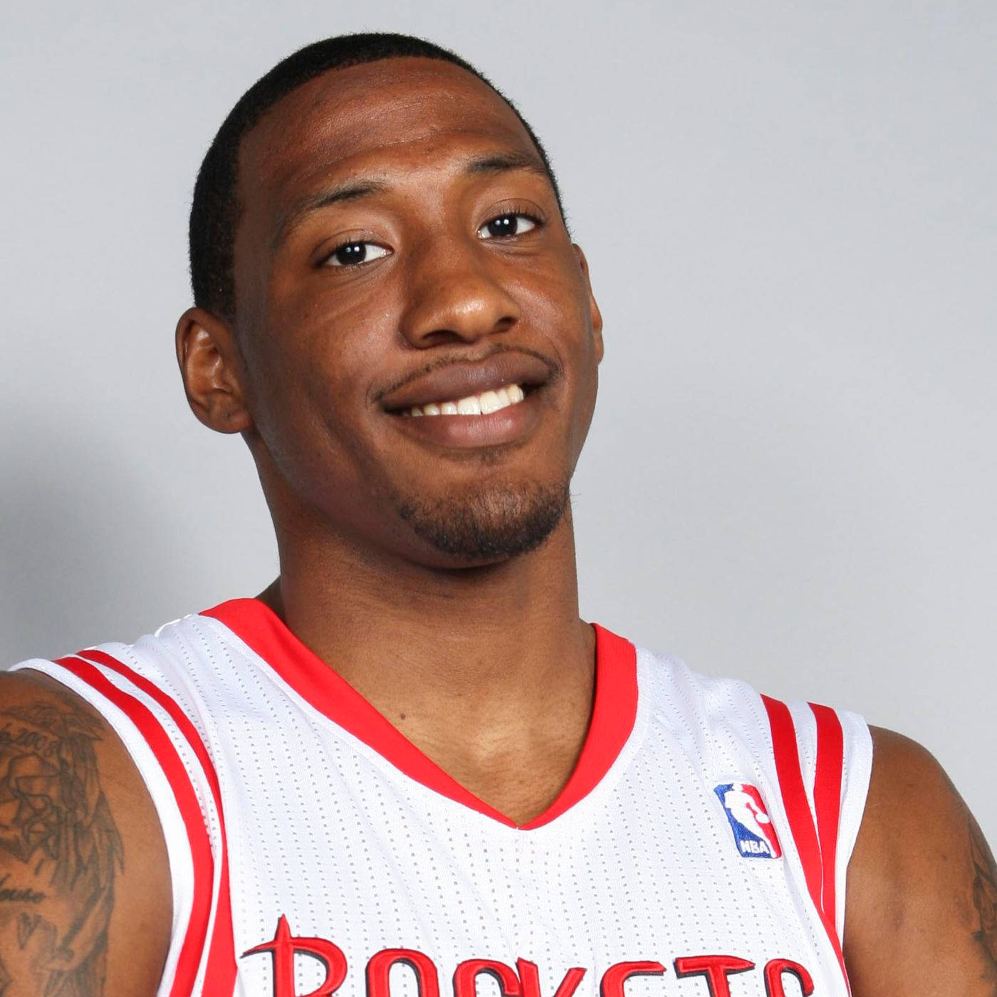 robert covington 桌布 壁紙 — 一位微笑的籃球運動員，身穿白色和紅色的火箭隊球衣，手臂上有刺青，背景為純灰色。