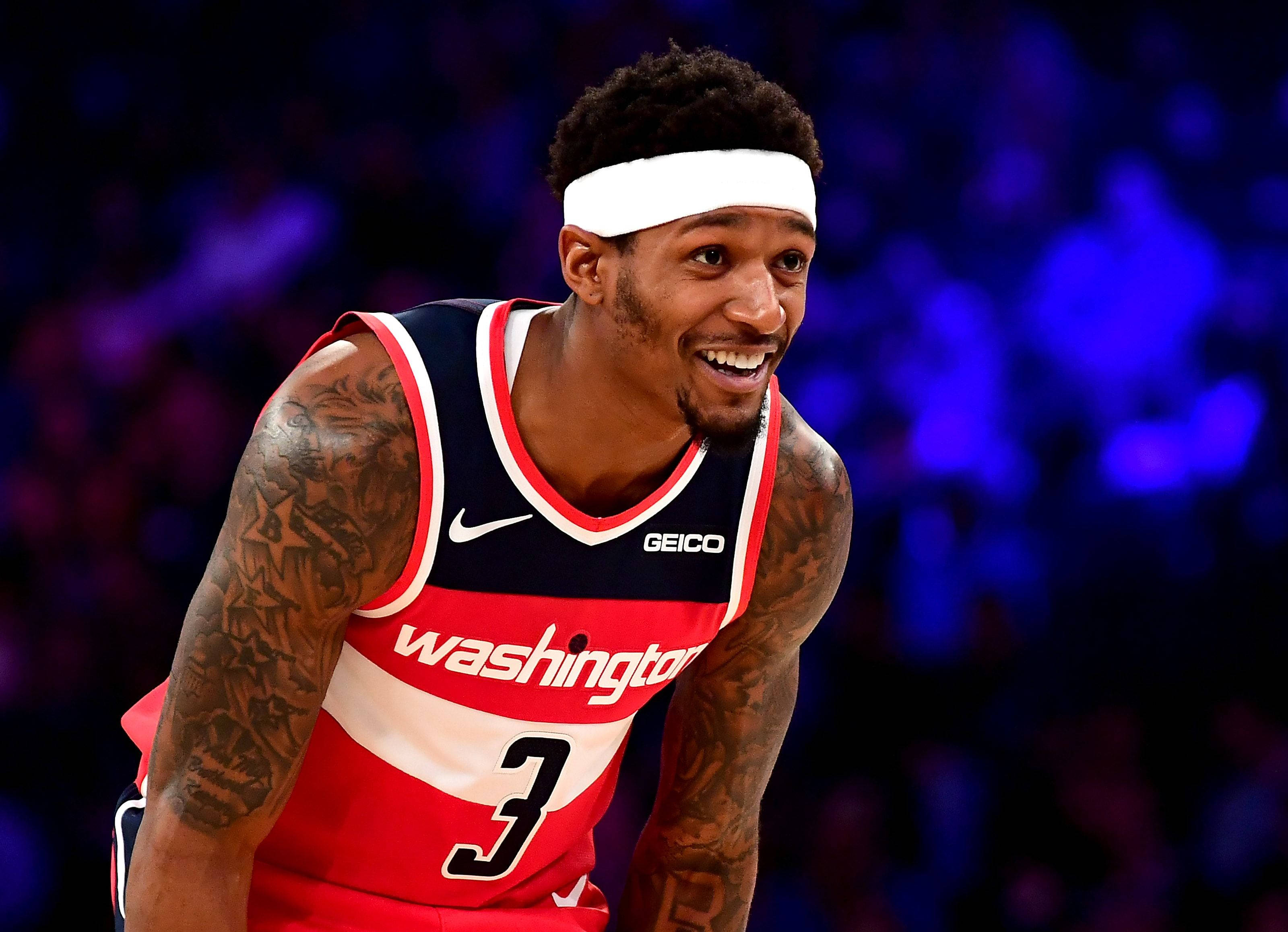 Bradley Beal 桌布 壁紙 — 一位微笑的籃球運動員，身穿紅白藍華盛頓球衣，有刺青和白色頭帶，背景是模糊的藍色球場。