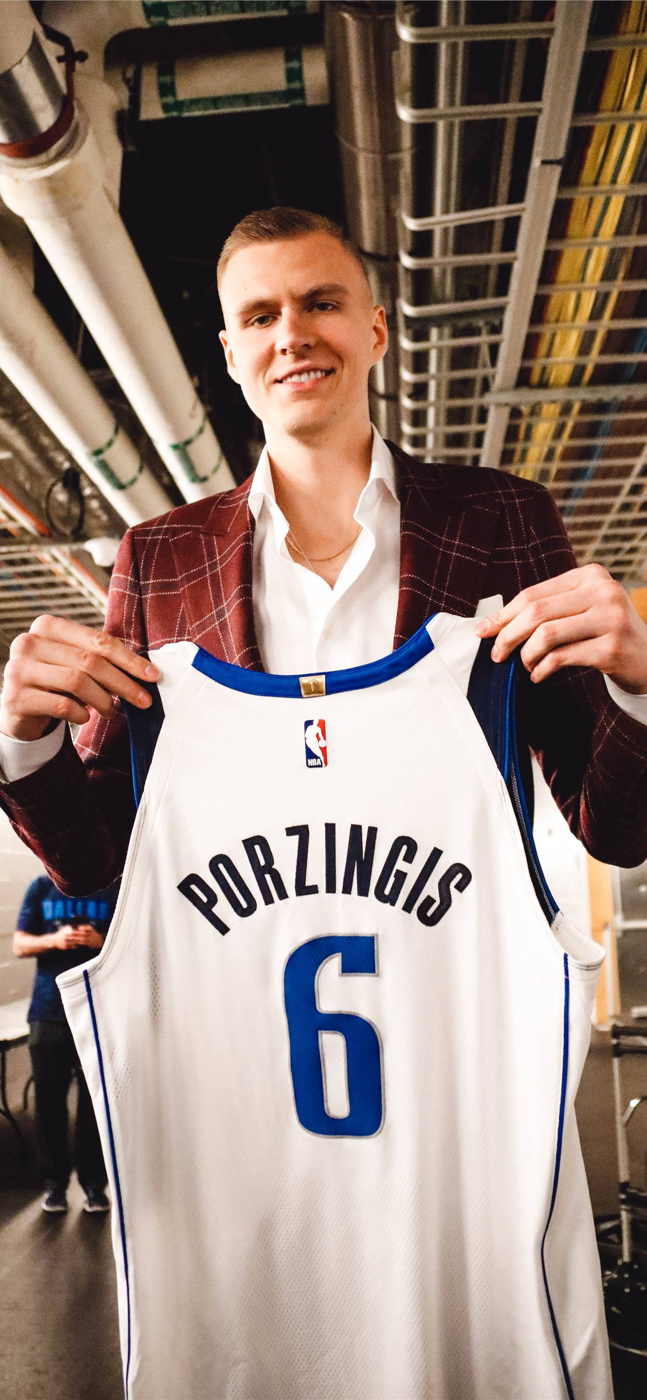 Kristaps Porzingis 桌布 壁紙 — 一位微笑的籃球運動員穿著格子西裝外套，在體育館自豪地舉起他的白色6號Porzingis球衣。