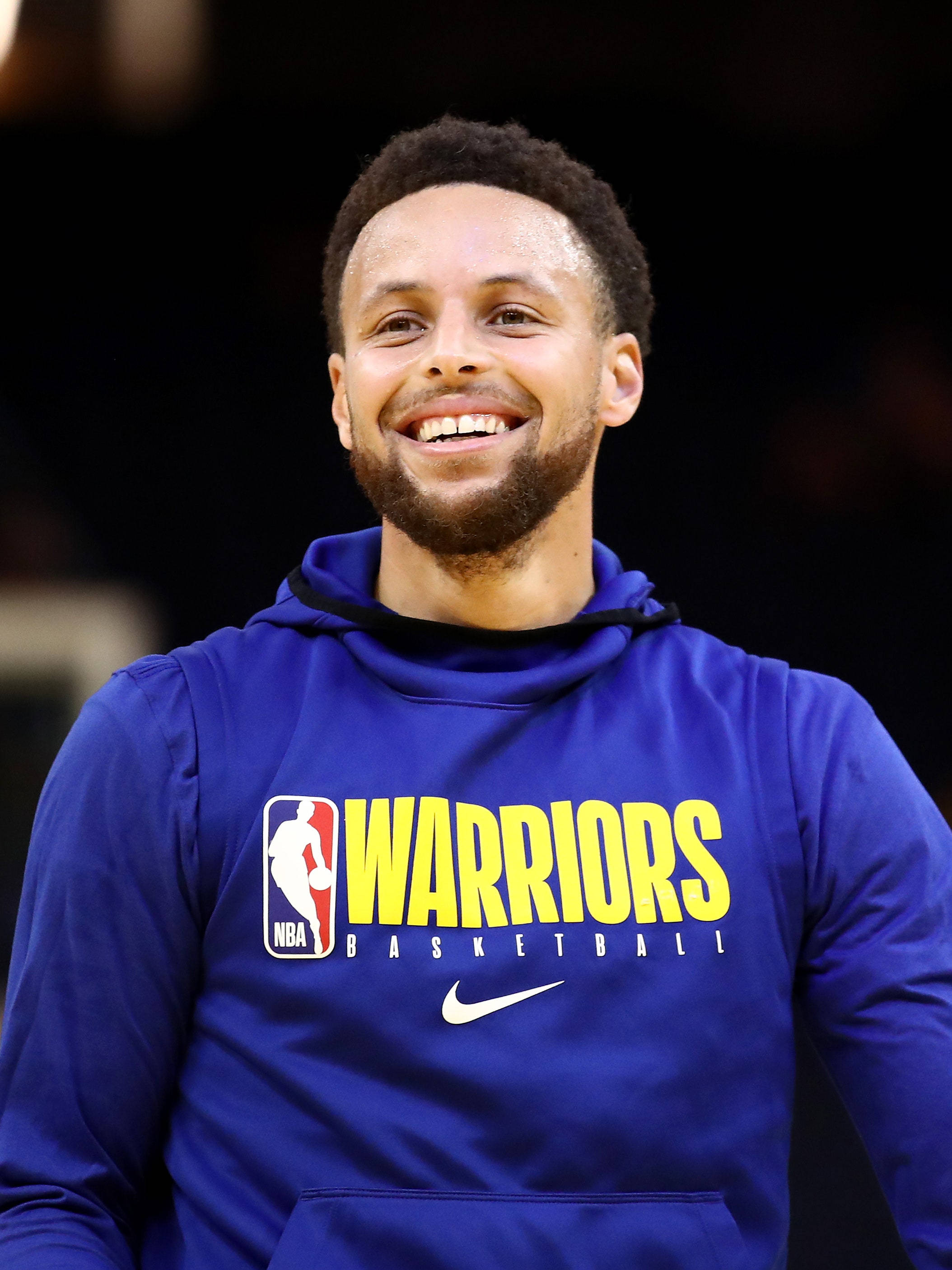 籃球 桌布 壁紙 — 一位微笑的籃球運動員穿著藍色Warriors連帽衫，站在模糊的深色背景前。