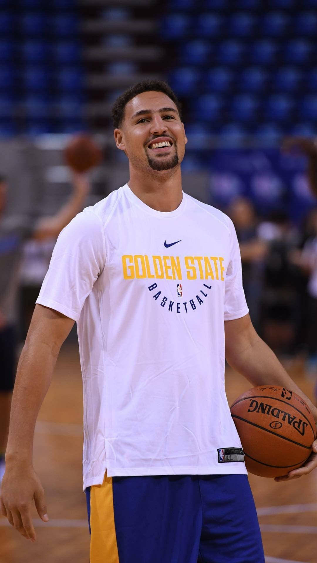 Thompson 桌布 壁紙 — 一位微笑的籃球運動員穿著金州勇士隊裝備，在硬木球場上拿著Spalding籃球，背景是藍色座位。