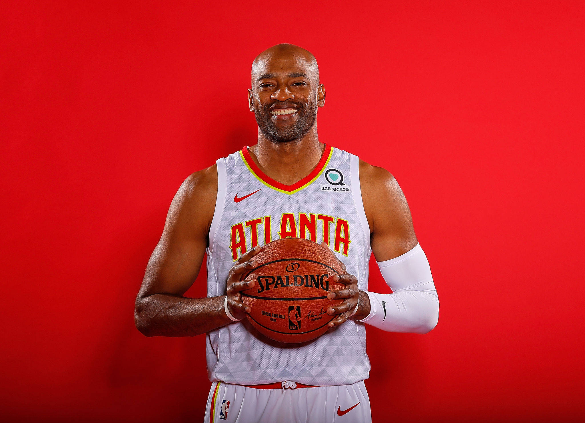 NBA 桌布 壁紙 — 一位微笑的籃球運動員身穿亞特蘭大球衣，在純紅色攝影棚背景前拿著一顆Spalding籃球。