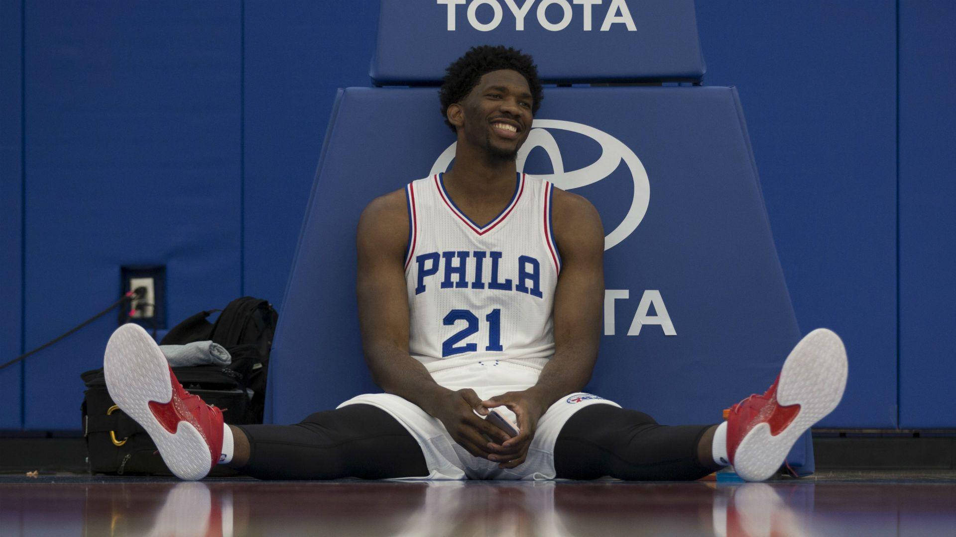 Embiid 桌布 壁紙 — 一位微笑的籃球運動員身穿白色Phila球衣，盤腿坐在拋光的地板上，腳穿紅白運動鞋。