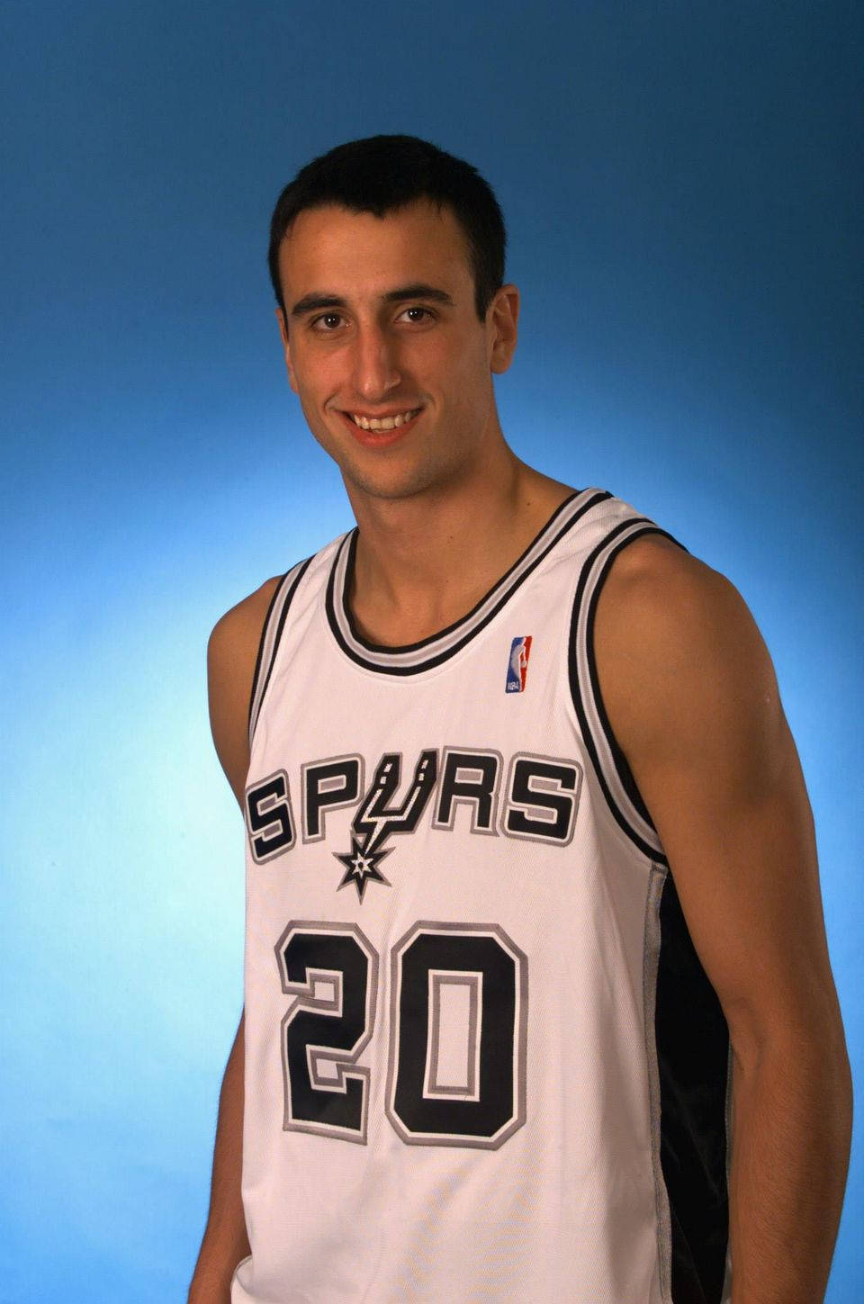 Manu 桌布 壁紙 — 一位微笑的籃球運動員身穿白色馬刺隊球衣，背號20，站在漸變藍色攝影棚背景前。