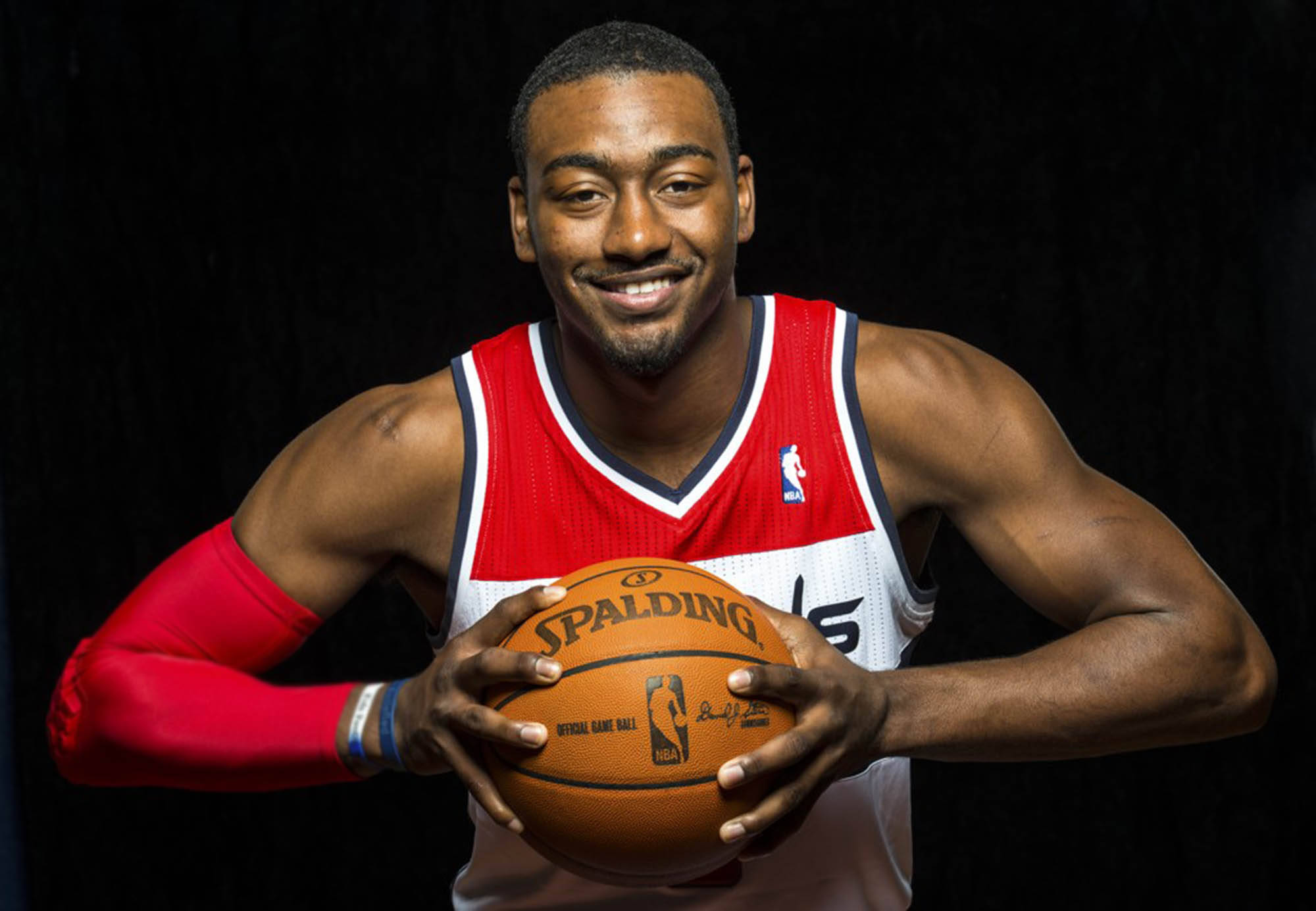 John Wall 桌布 壁紙 — 一位微笑的籃球運動員身穿紅白球衣，在黑色背景前拿著一個 Spalding 籃球。