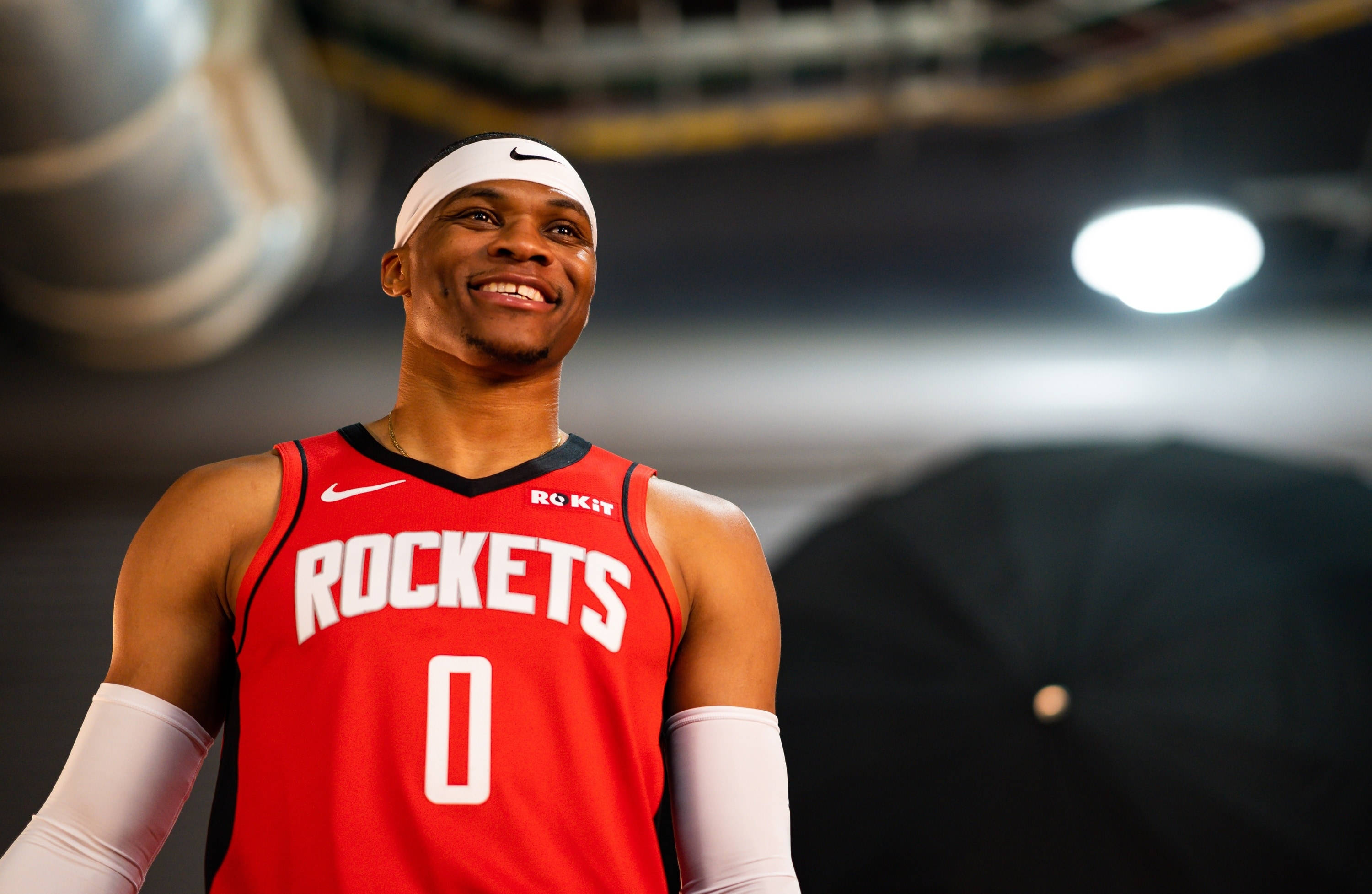 Westbrook 桌布 壁紙 — 一位微笑的籃球運動員身穿紅色火箭隊球衣，號碼零，戴著白色頭帶，自信地站在燈光昏暗的球場上。