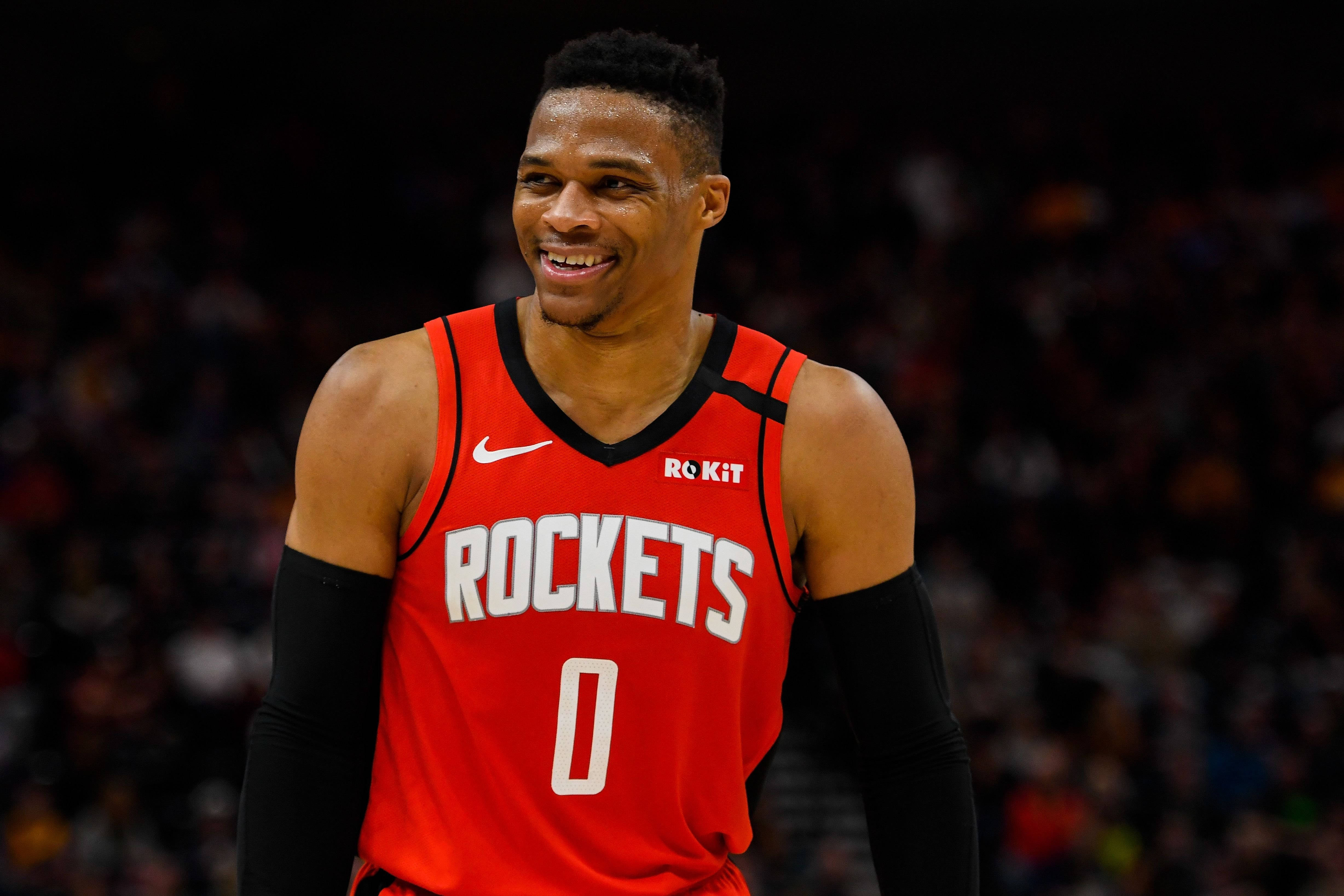 Westbrook 桌布 壁紙 — 一位微笑的籃球運動員身穿紅色火箭隊球衣，號碼為零，站在明亮的場館燈光下的球場上。