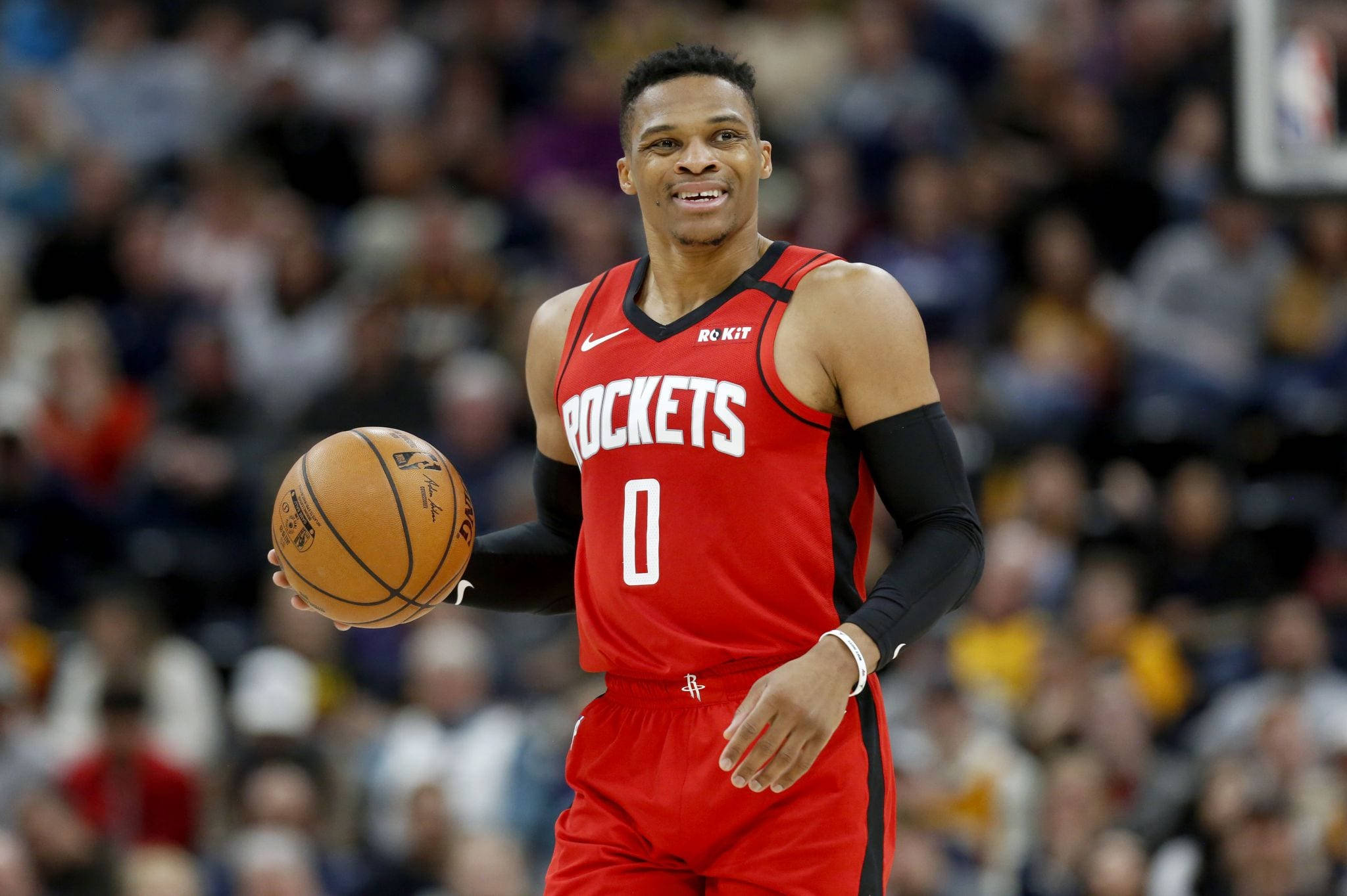 Westbrook 桌布 壁紙 — 一位微笑的籃球運動員身穿紅色火箭隊球衣，在比賽中持球，身後是模糊的人群。