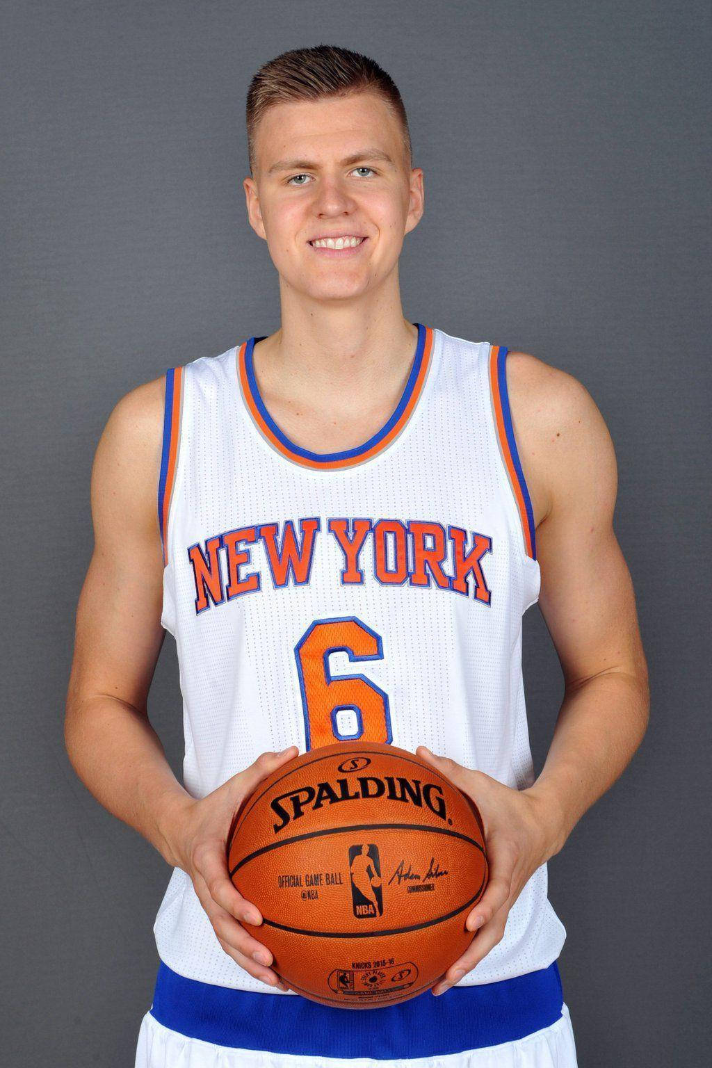 Kristaps Porzingis 桌布 壁紙 — 一位微笑的籃球運動員身穿紐約尼克隊球衣，在純灰色工作室背景前拿著一個Spalding籃球。