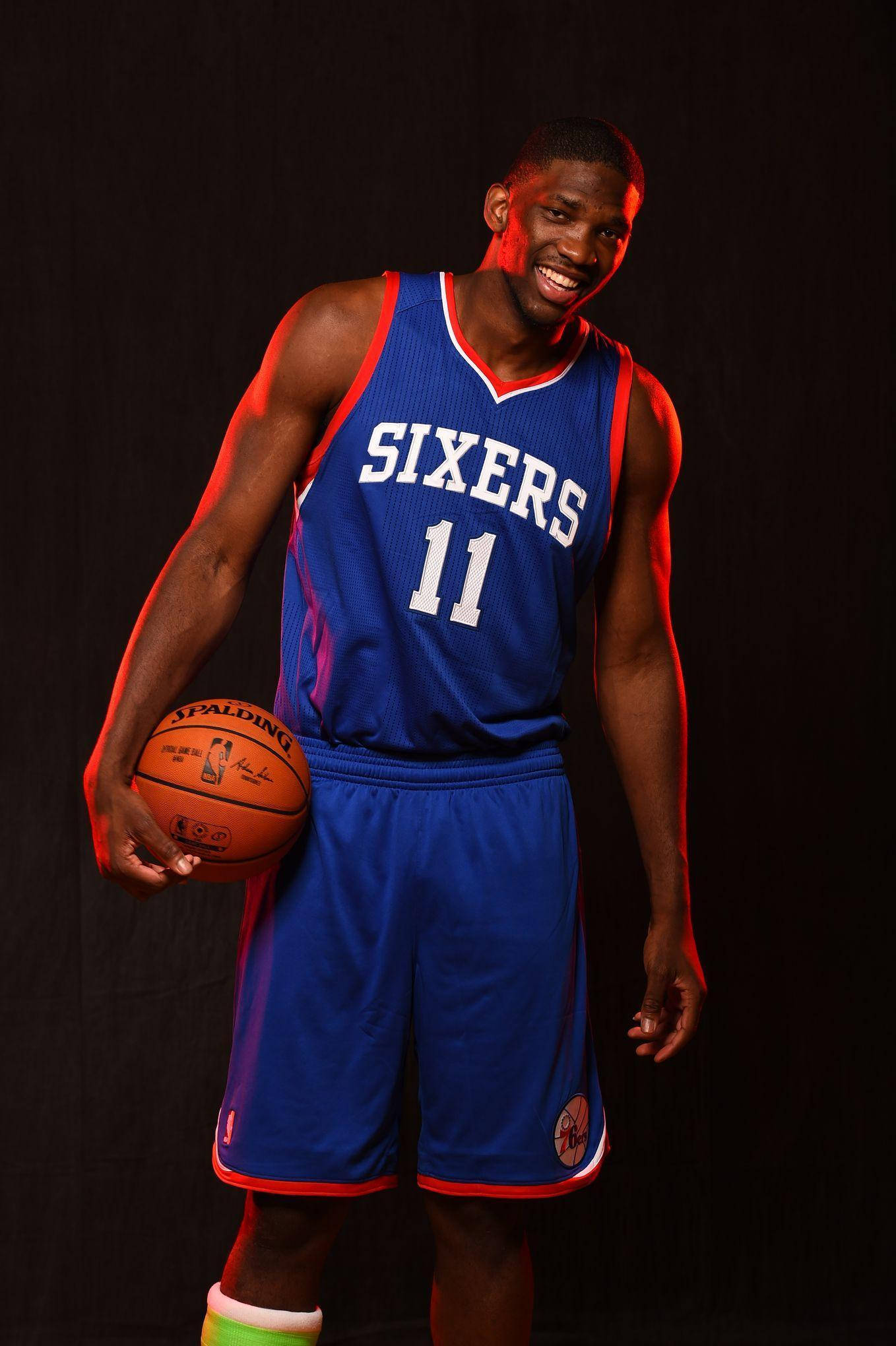 Embiid 桌布 壁紙 — 一位微笑的籃球運動員身穿藍色76人球衣，在帶有紅色燈光效果的黑暗背景下手持Spalding籃球。