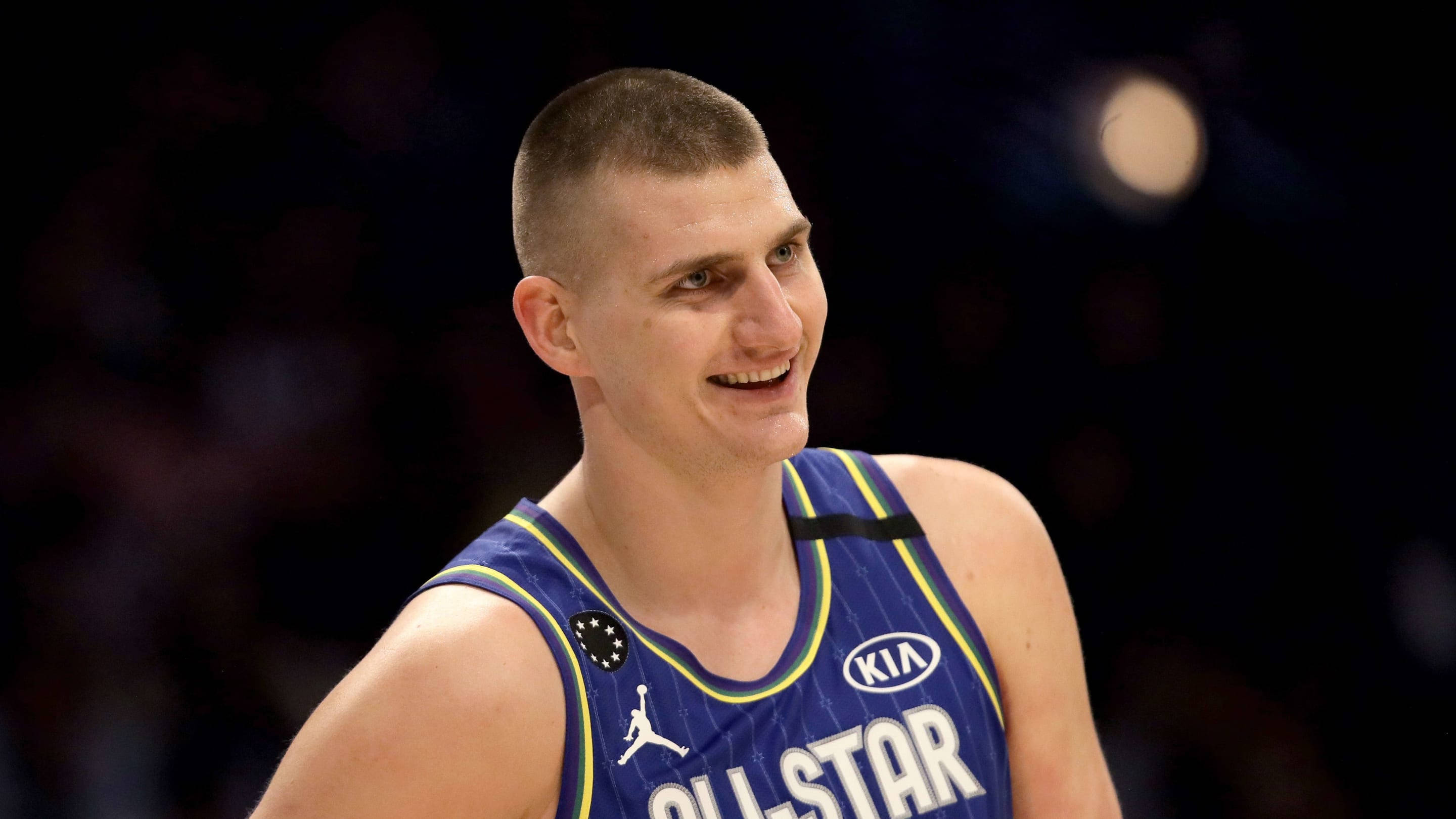 Jokic 桌布 壁紙 — 一位微笑的籃球運動員身穿藍色全明星球衣，帶有黃色飾邊和Kia標誌，在黑暗模糊的球場背景下看向鏡頭外。