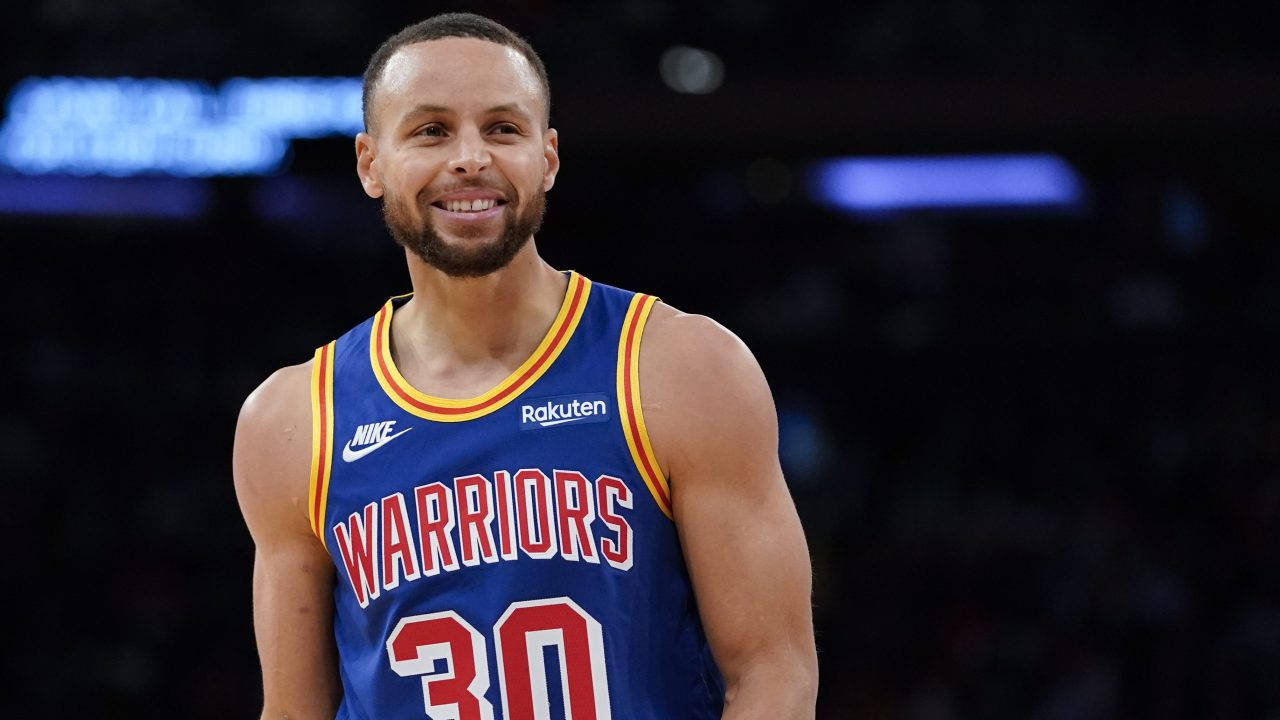 NBA 桌布 壁紙 — 一位微笑的籃球運動員身穿藍色勇士隊球衣，背號30，在明亮的場館燈光下自信地站在球場上。