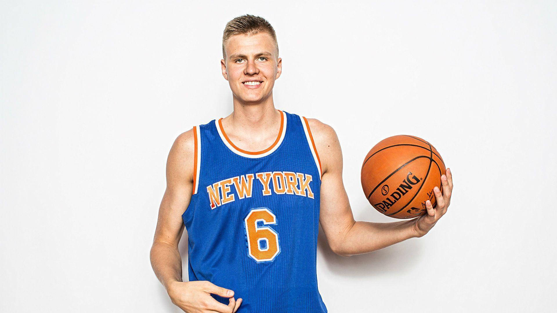 Kristaps Porzingis 桌布 壁紙 — 一位微笑的籃球運動員身穿藍色紐約球衣，在純白色背景前手持橙色 Spalding 籃球。
