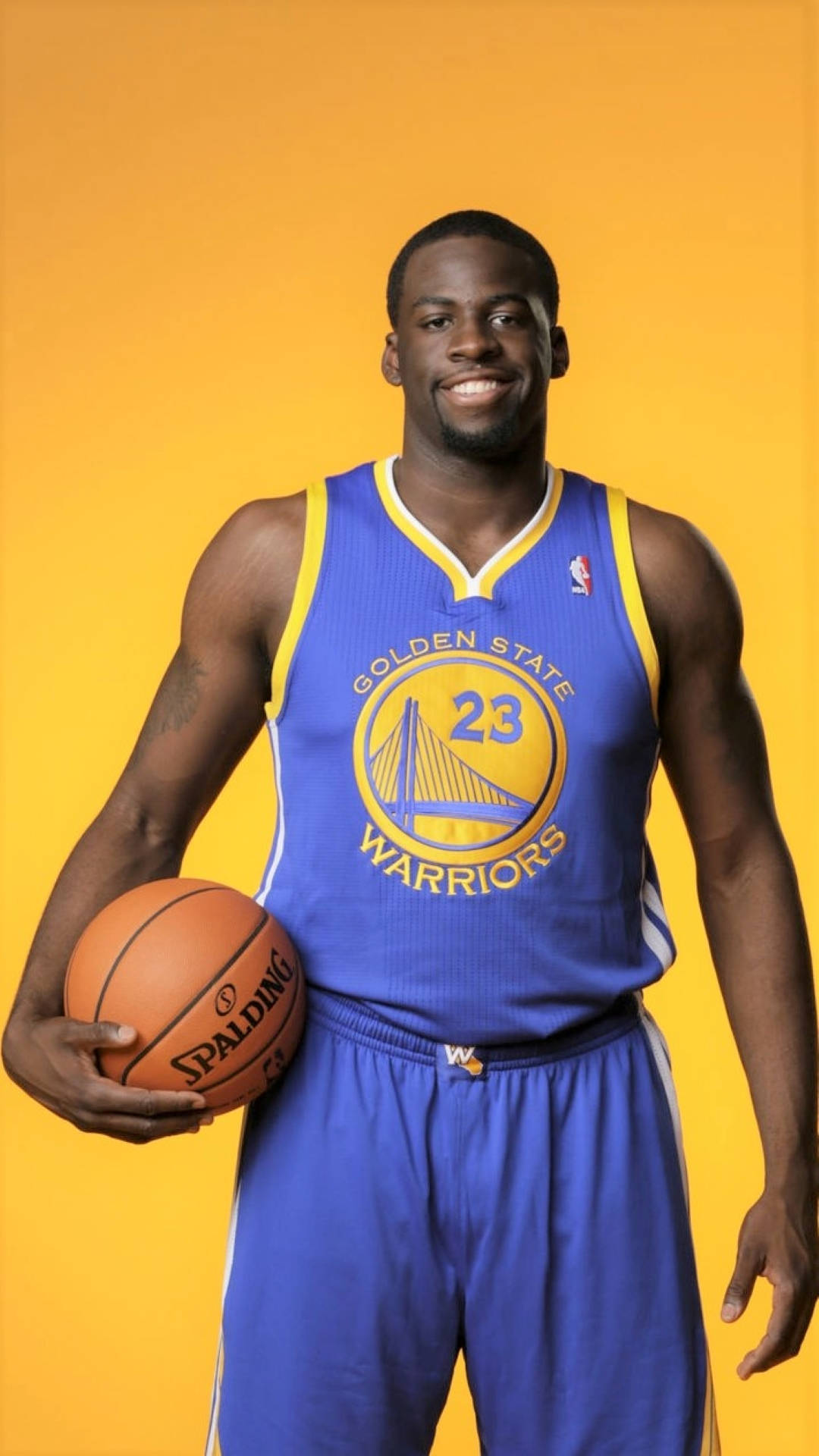 Draymond Green 桌布 壁紙 — 一位微笑的籃球運動員身穿金州勇士隊球衣，在純黃色背景前拿著一個Spalding籃球。