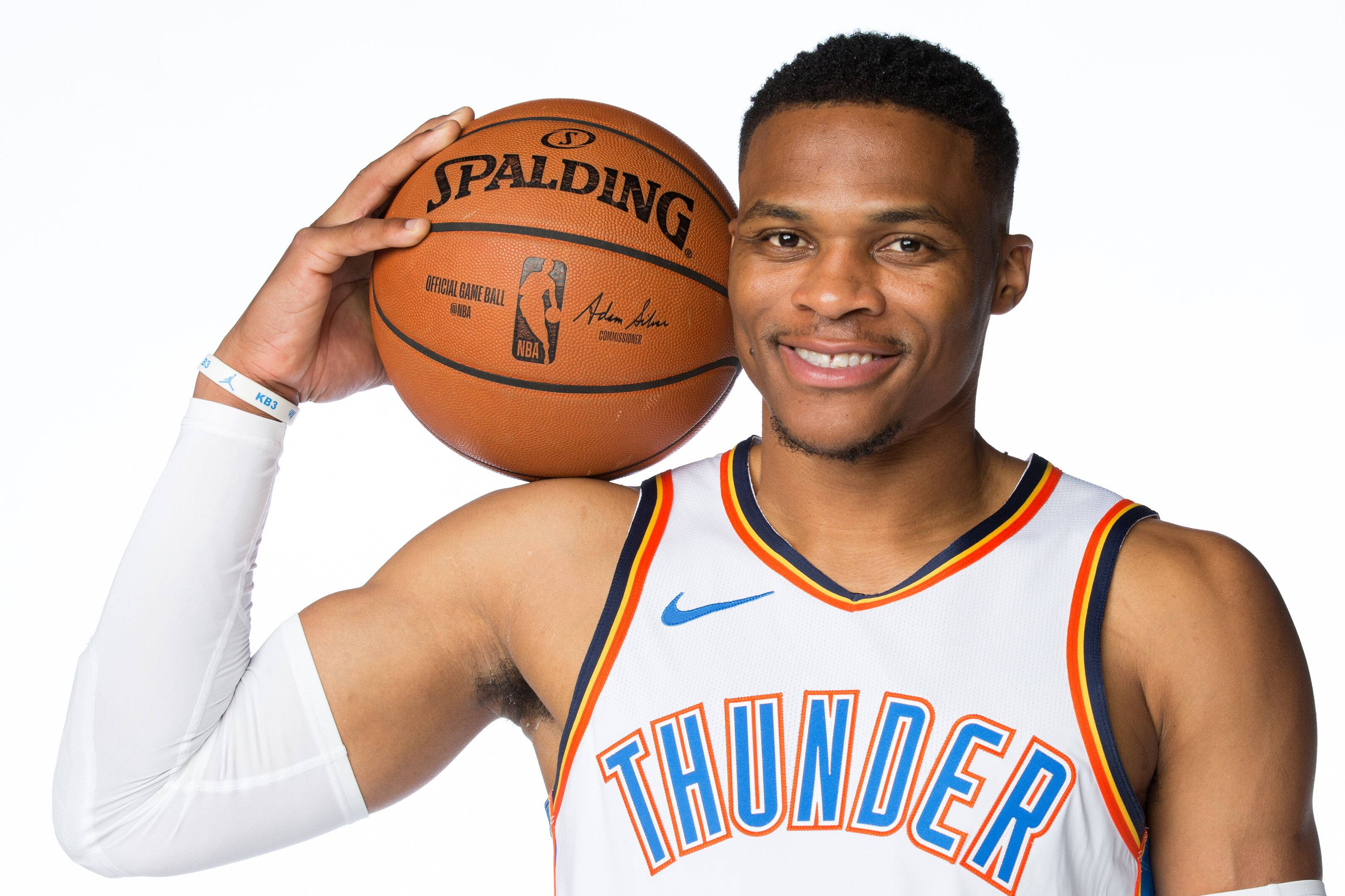 Westbrook 桌布 壁紙 — 一位微笑的籃球運動員身穿雷霆隊球衣，在白色背景前手持 Spalding NBA 比賽用球。
