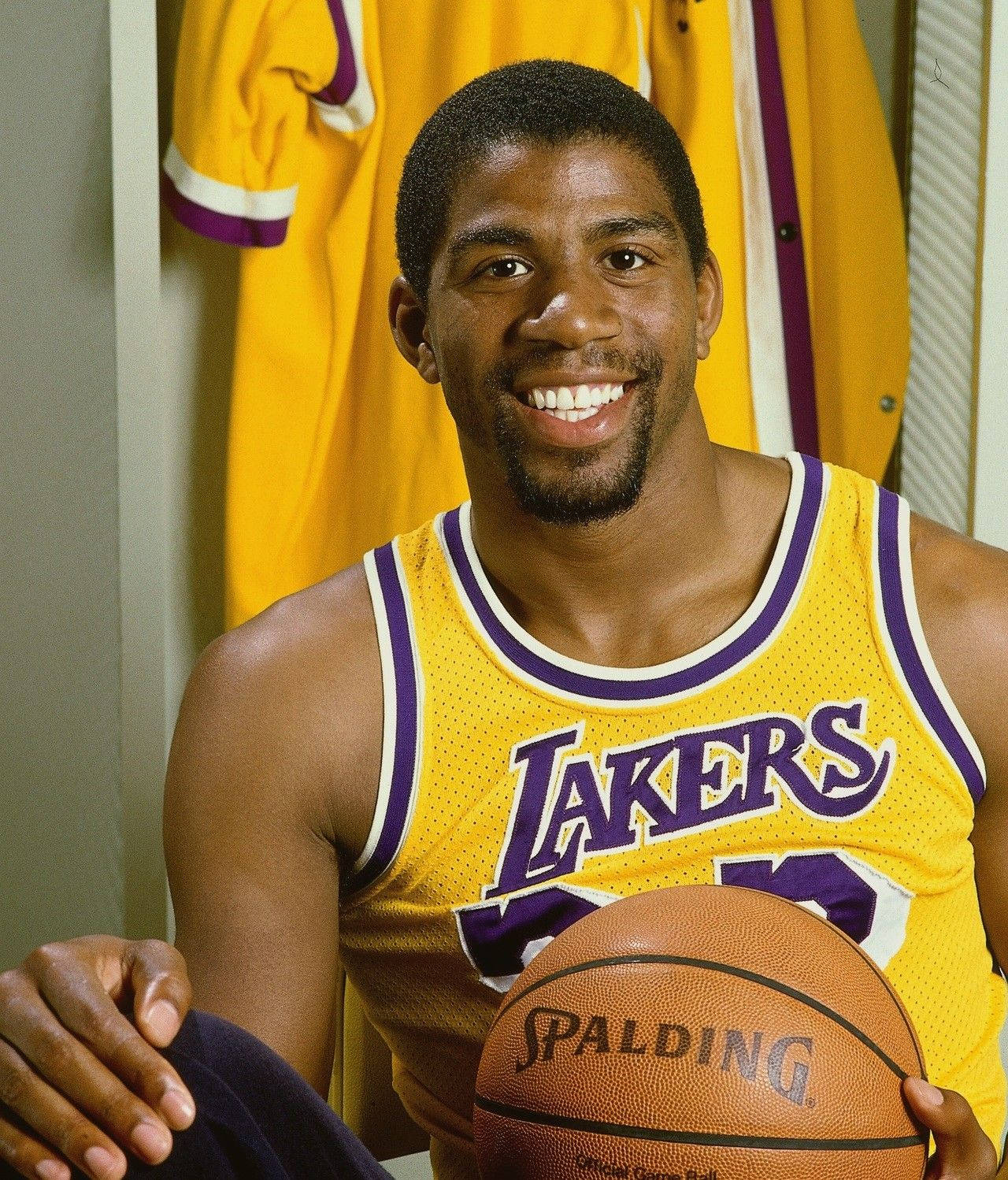 Magic Johnson 桌布 壁紙 — 一位微笑的籃球運動員身穿黃色湖人隊球衣，在更衣室裡拿著一個 Spalding 籃球，身後掛著球衣。