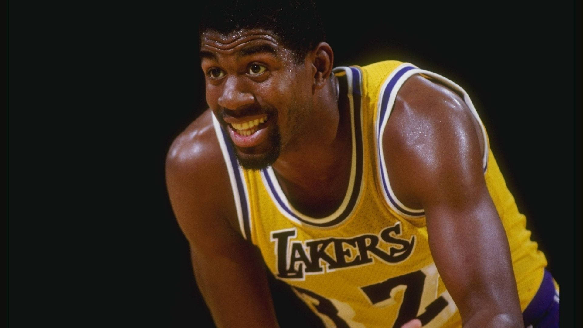 Magic Johnson 桌布 壁紙 — 一位微笑的籃球運動員身穿黃色湖人隊球衣，在黑色背景前向前傾身。