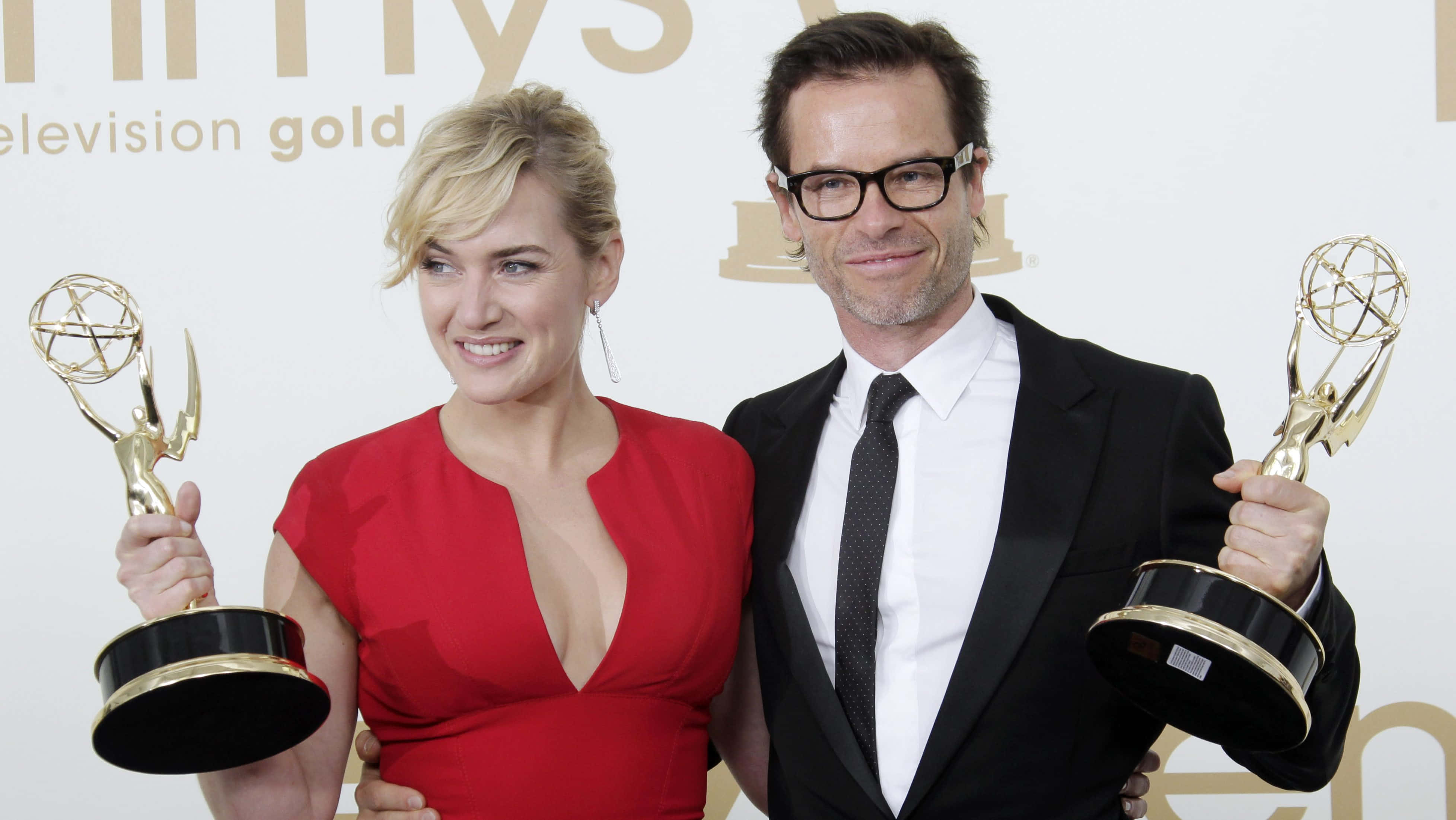 Guy Pearce 桌布 壁紙 — 一位微笑的紅裙女士和一位黑西裝男士各自手持金色艾美獎獎座，背景為白色並有金色文字。