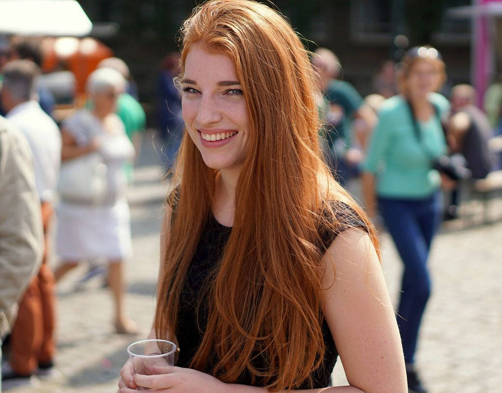 smiling 桌布 壁紙 — 一位微笑的紅髮女性，長髮飄逸，在繁忙的戶外人群中手持飲品。