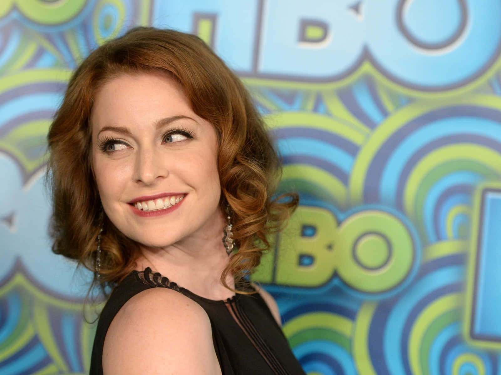 Esmé Bianco 桌布 壁紙 — 一位微笑的紅髮女性戴著垂墜耳環，在色彩繽紛的漩渦背景和「HBO」標誌前擺姿勢。