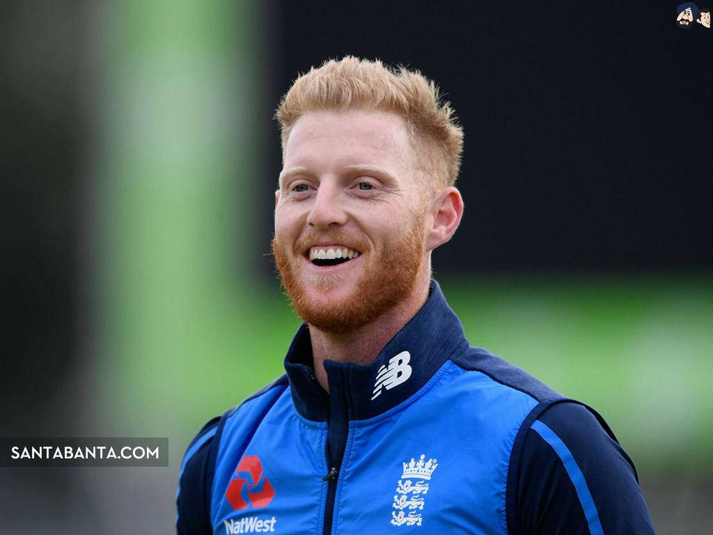 Ben Stokes 桌布 壁紙 — 一位微笑的紅髮板球運動員穿著藍色英格蘭訓練外套，在模糊的綠色背景前開懷大笑。