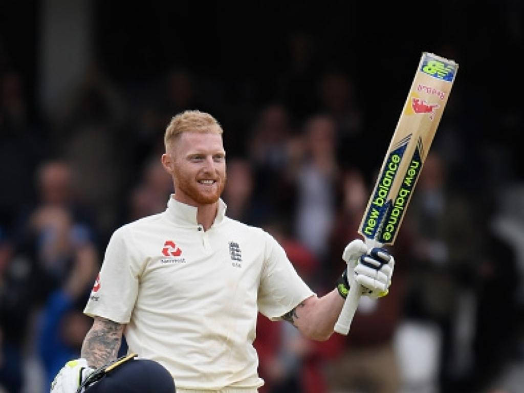 Ben Stokes 桌布 壁紙 — 一位微笑的紅髮板球運動員身穿白色制服，勝利地高舉球棒，向歡呼的球迷致意。