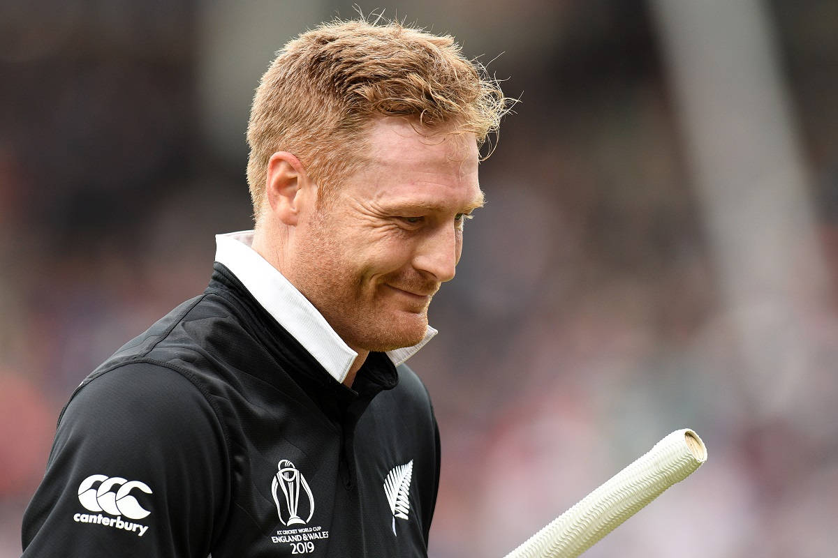 martin guptill 桌布 壁紙 — 一位微笑的紐西蘭板球運動員身穿黑色球衣，在模糊的體育場人群前手持白色球棒。