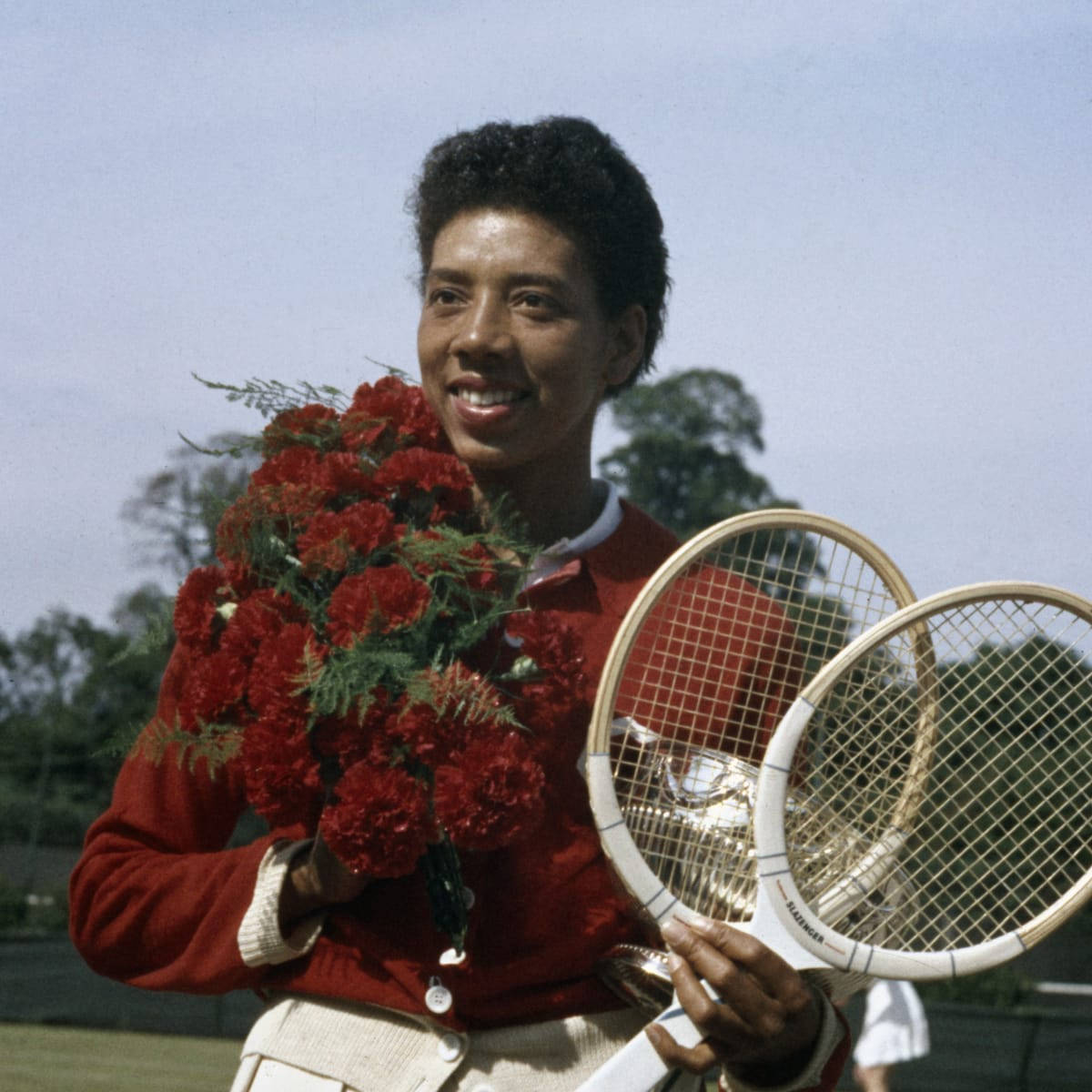 Tennis 桌布 壁紙 — 一位微笑的網球運動員穿著紅色毛衣，在戶外手持兩把復古球拍和一束紅色康乃馨。