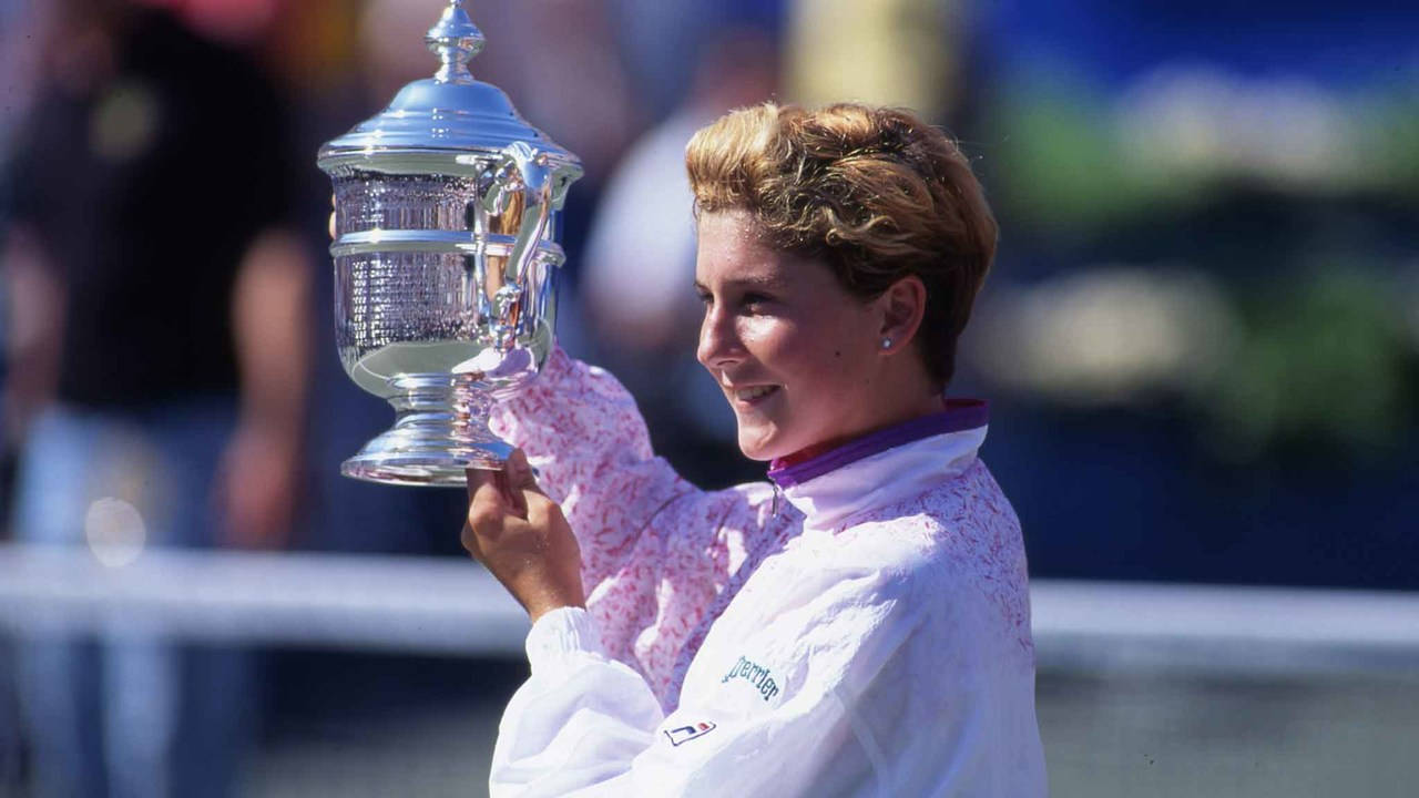 Monica Seles 桌布 壁紙 — 一位微笑的網球運動員身穿白色外套，在明亮的陽光下自豪地舉起一個大型銀色獎盃。