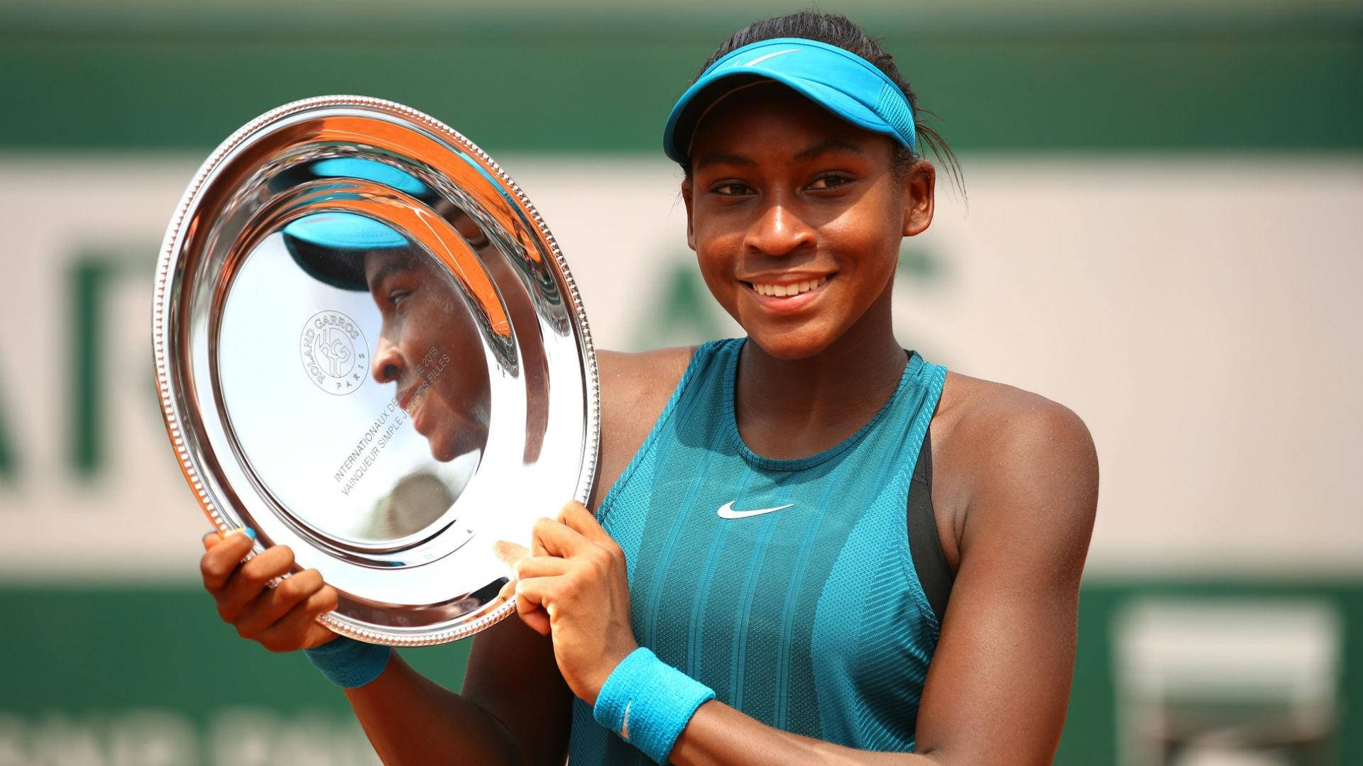 Coco Gauff 桌布 壁紙 — 一位微笑的網球運動員身穿藍色衣服，在紅土球場上自豪地舉起一個大型銀色獎盃。