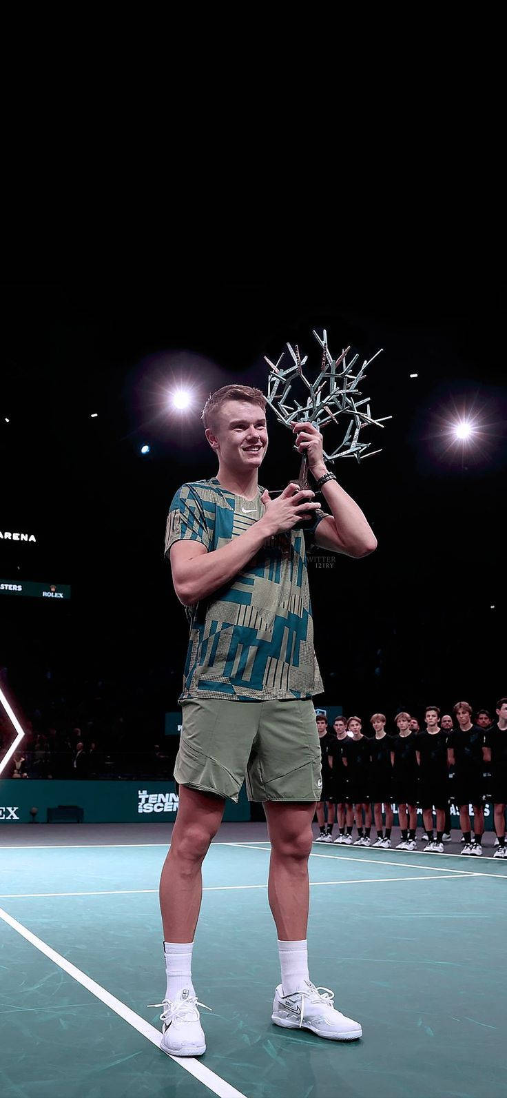 Champion 桌布 壁紙 — 一位微笑的網球選手在光線明亮的室內球場上自豪地舉起一個獨特的銀色獎盃，背景有觀眾。