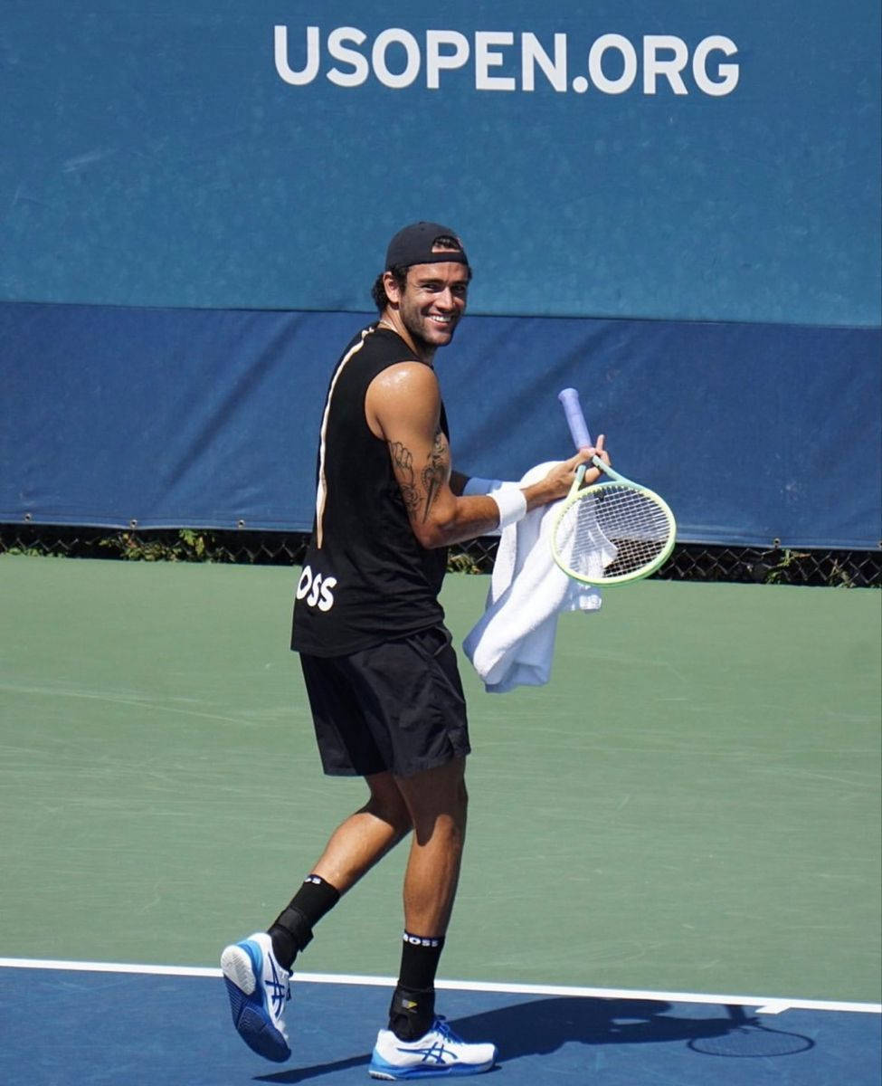 Tennis 桌布 壁紙 — 一位微笑的網球選手穿著黑色裝備，在明亮的陽光下拿著球拍和毛巾走在綠色球場上。