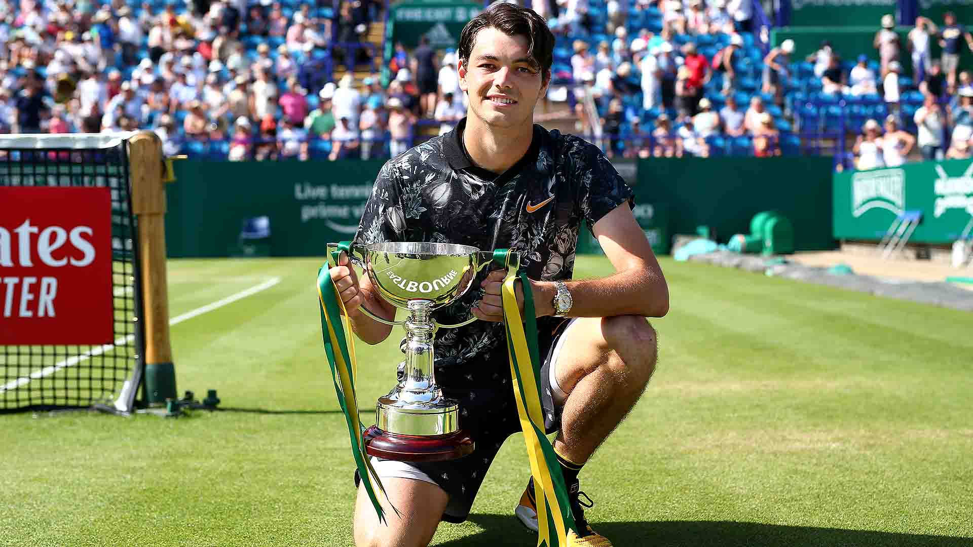Champion 桌布 壁紙 — 一位微笑的網球選手跪在草地場上，手持一個閃閃發光、繫有綠色和黃色絲帶的獎盃。